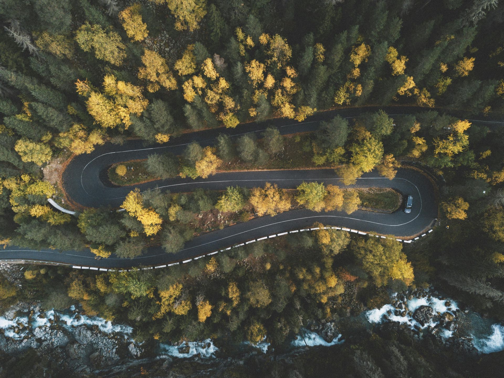 An aerial view of a winding asphalt road cutting through a dense autumn forest, with a river flowing along the bottom.