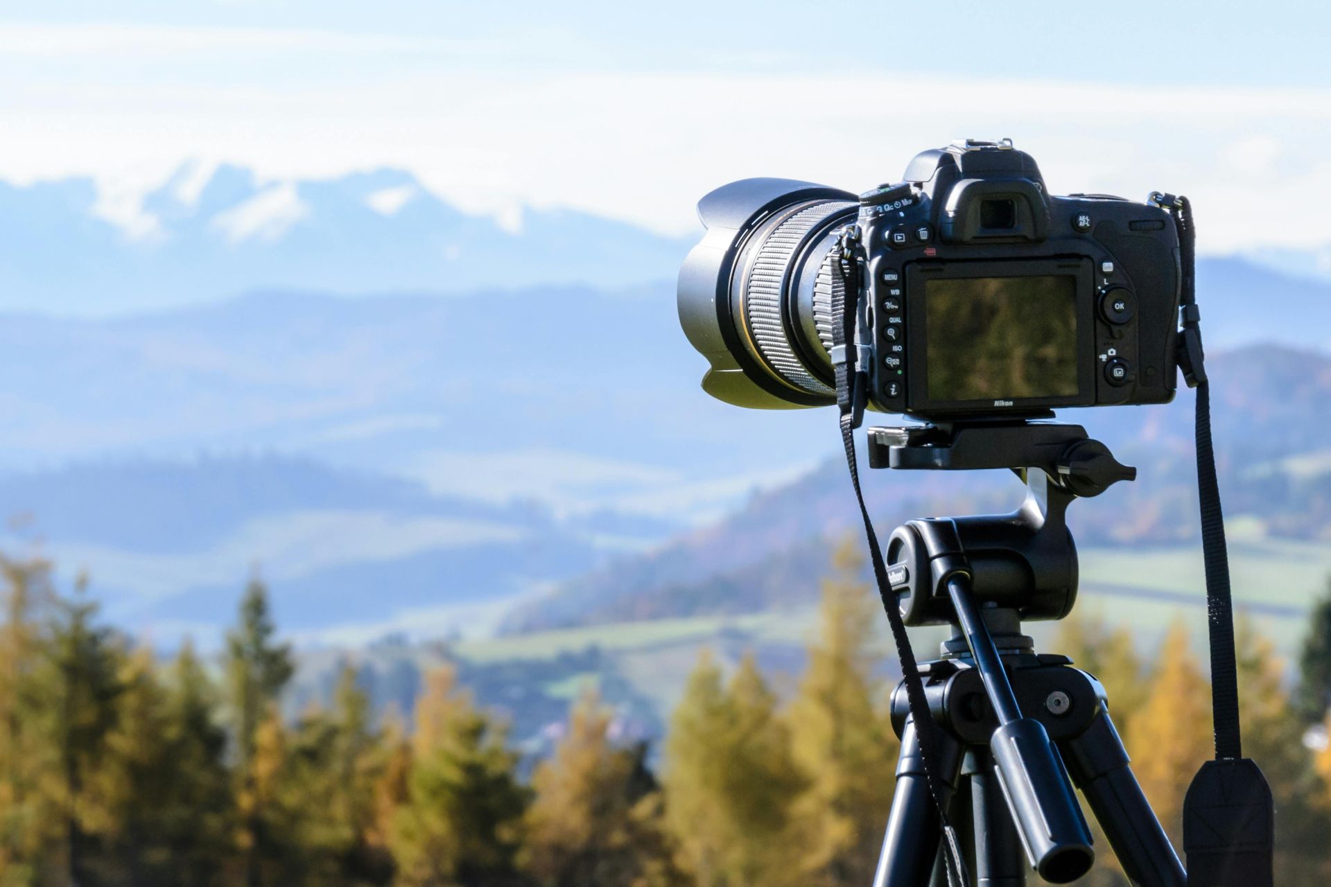 A professional camera on a tripod faces a scenic landscape of rolling hills and distant snow-capped mountains.