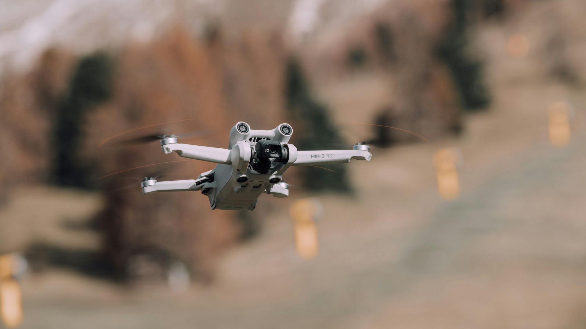 A grey drone hovers in mid-air against a blurred, out-of-focus forest background.