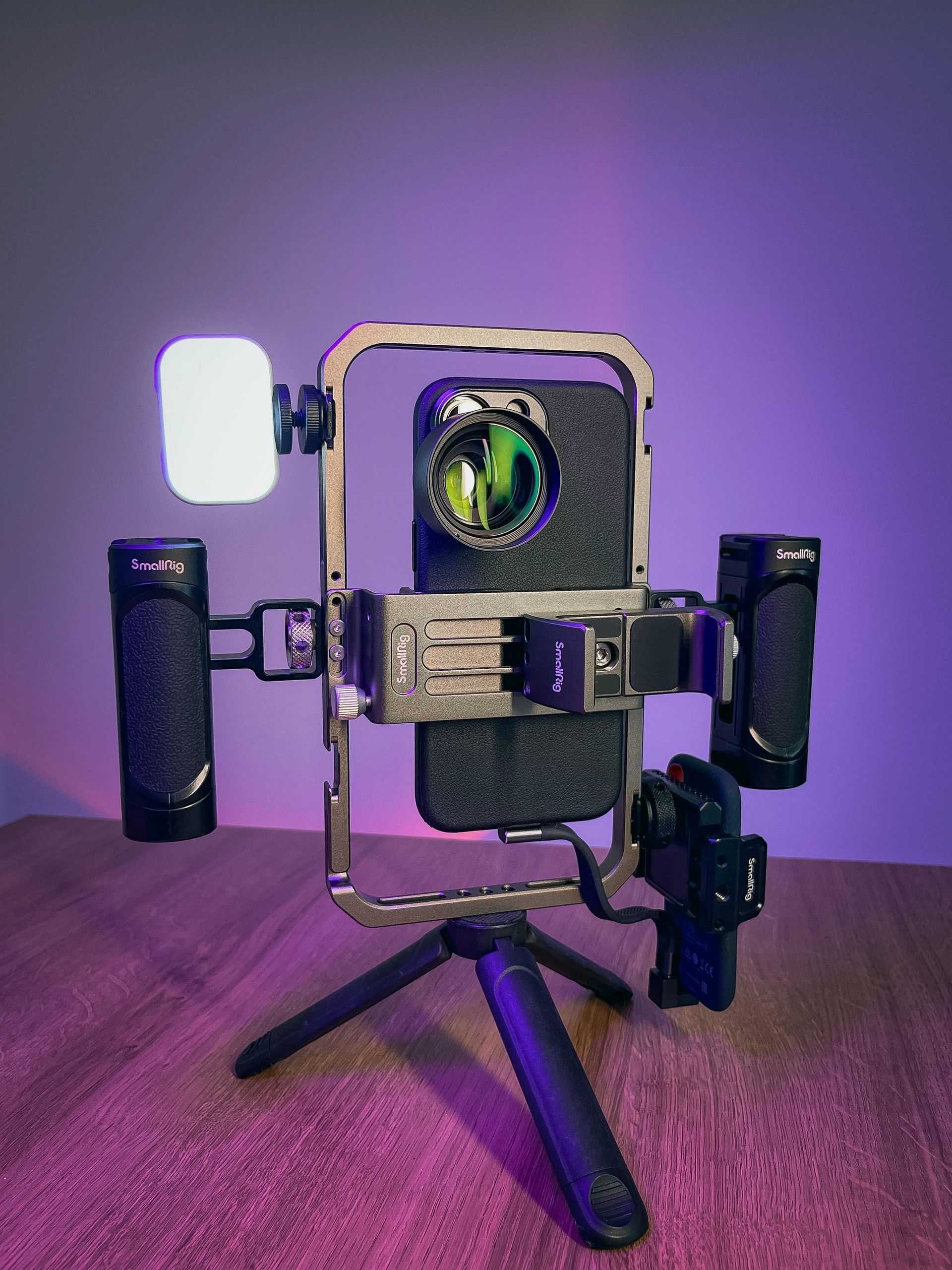 A smartphone in a metal rig with handles, a lens attachment, and a small LED light, mounted on a tripod against purple light.