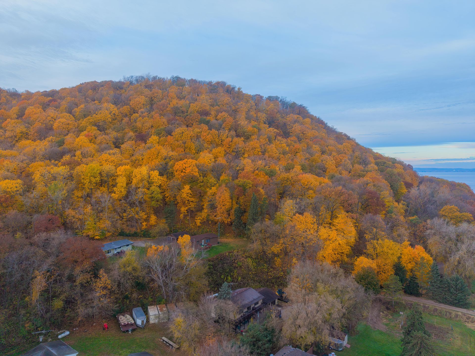 Autumn foliage covers a hill above buildings and a body of water under a blue sky.