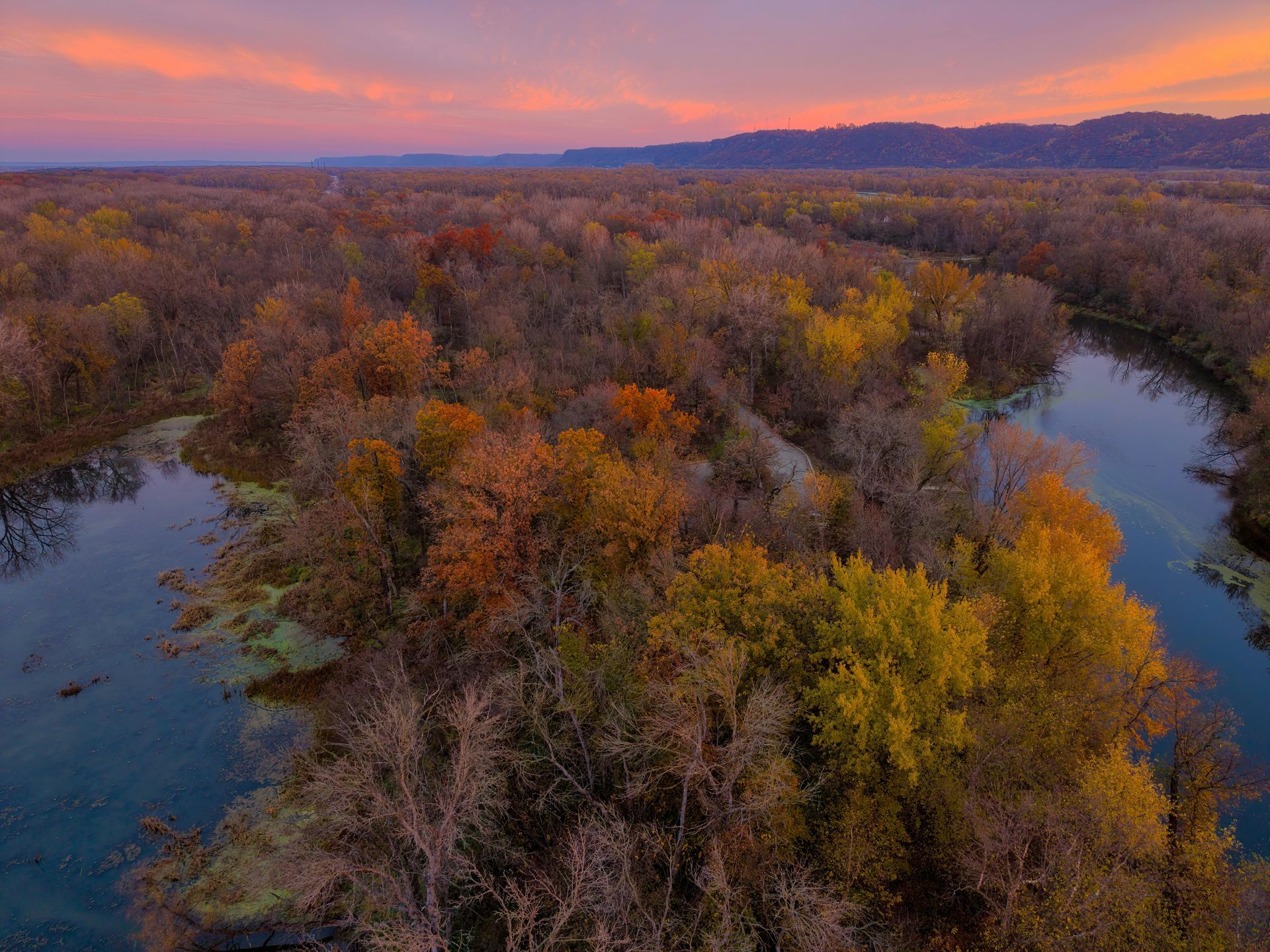 Aerial view of a colorful autumn forest with a winding river under a pink and orange sunset.