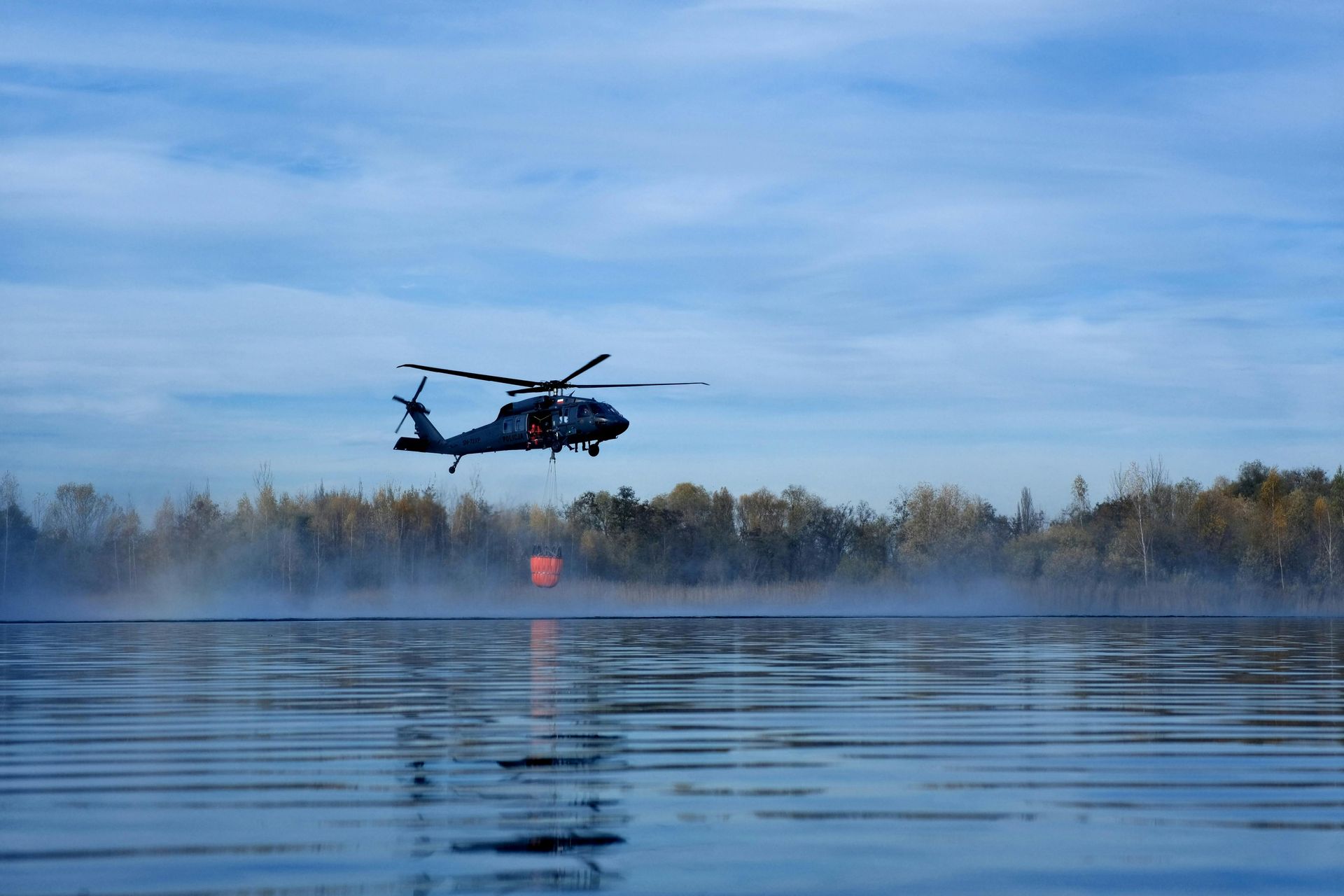 Helicopter over water refills a water bucket, likely for firefighting. Blue sky, water reflection, and trees.
