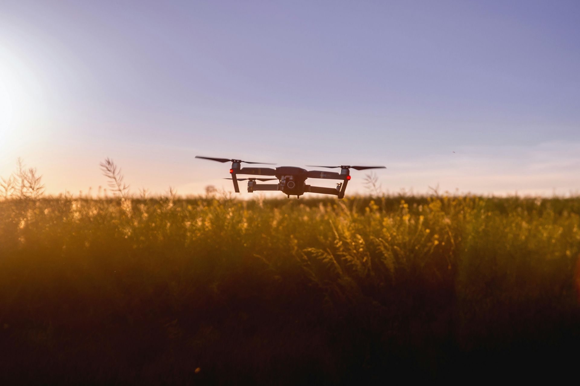 A drone hovers low over a field of golden grass during a sunset.