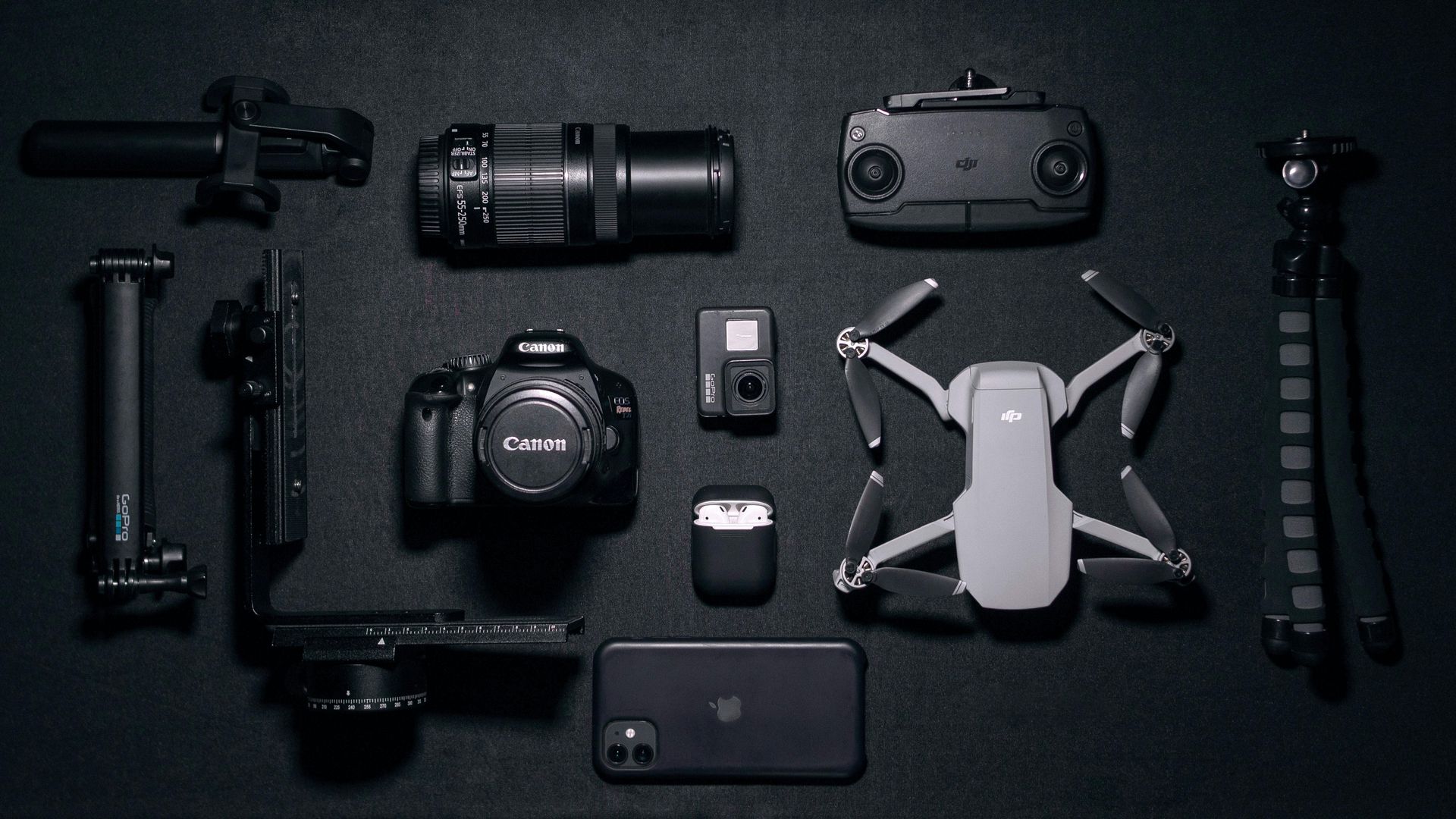 Flat lay on a dark surface featuring a camera, drone, controller, smartphone, tripod, and miscellaneous camera accessories.