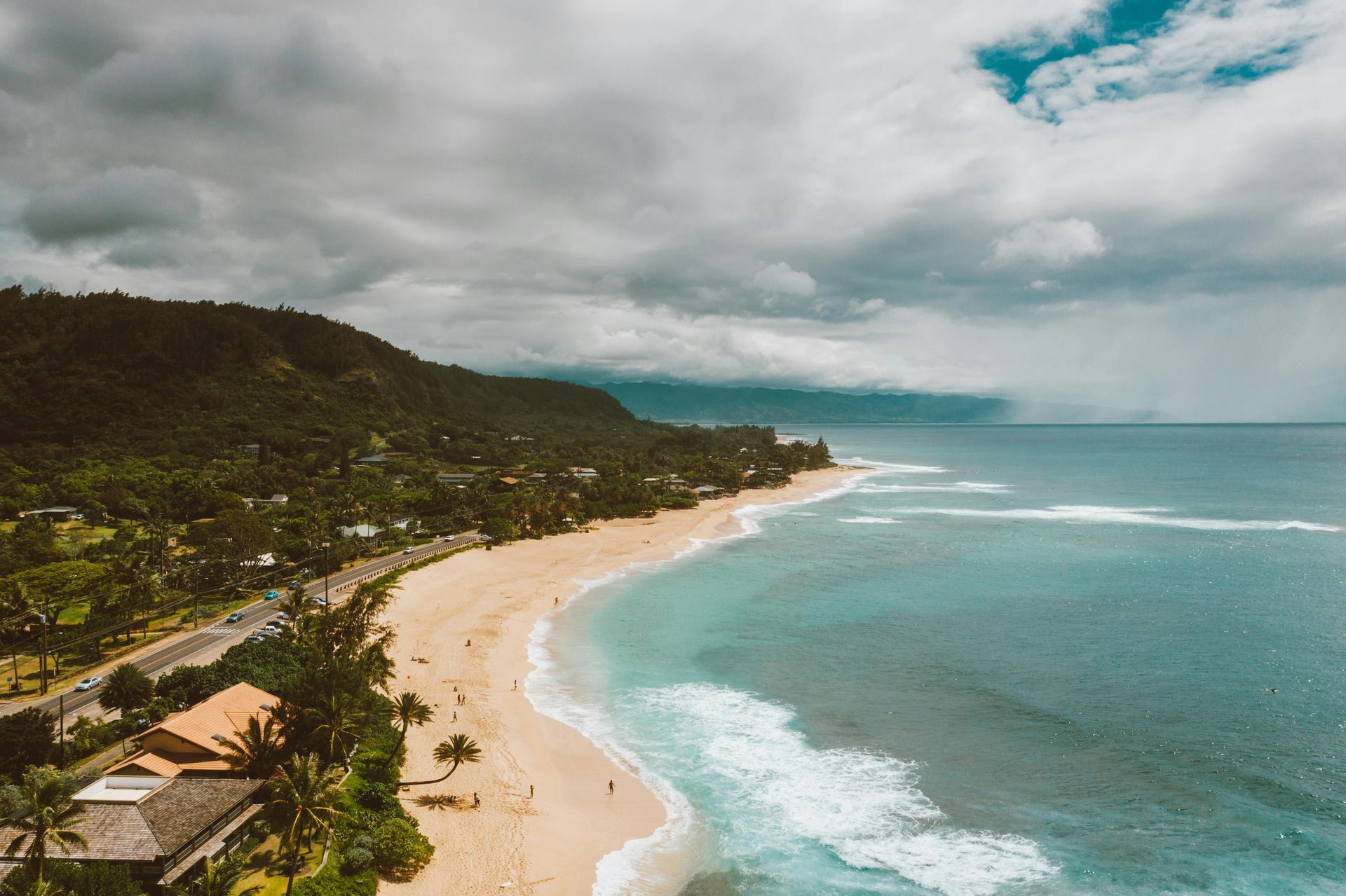 Beach with golden sand, turquoise water, green mountains under a cloudy sky.