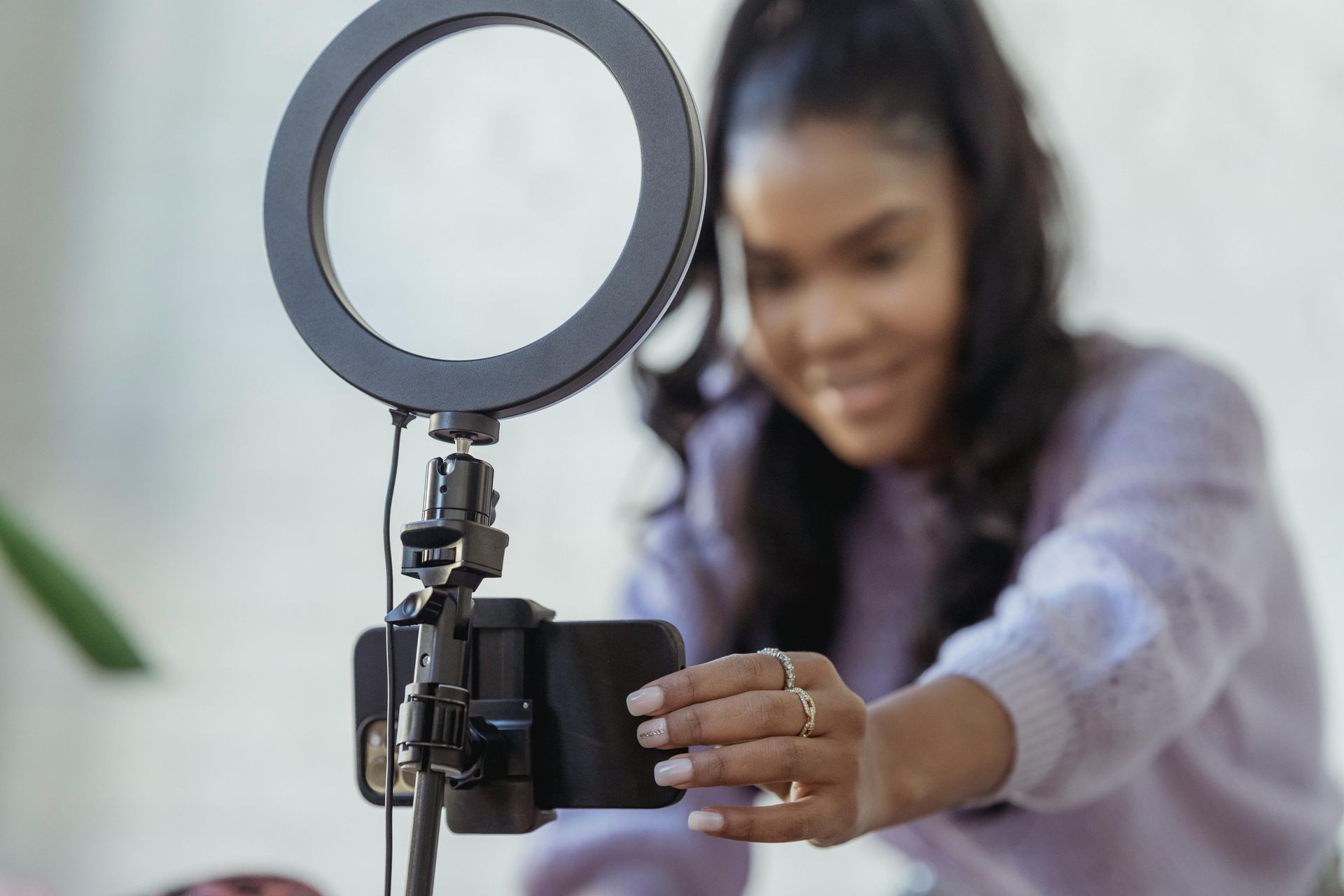 A person in a lavender sweater adjusting a smartphone mounted on a stand with a circular ring light in a bright room.