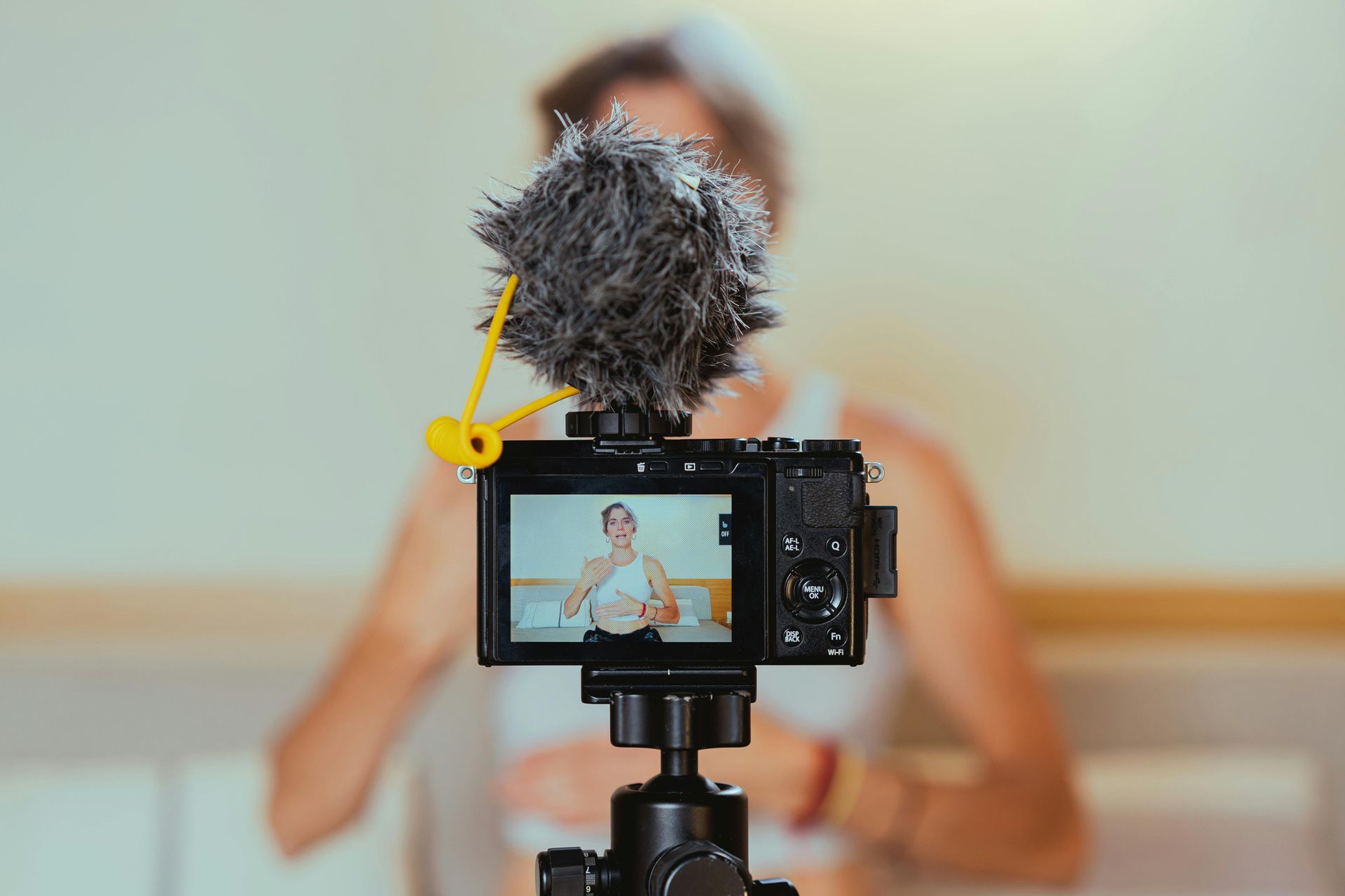 A camera with a fuzzy microphone windscreen mounted on a tripod, showing a person on the screen recording in a room.