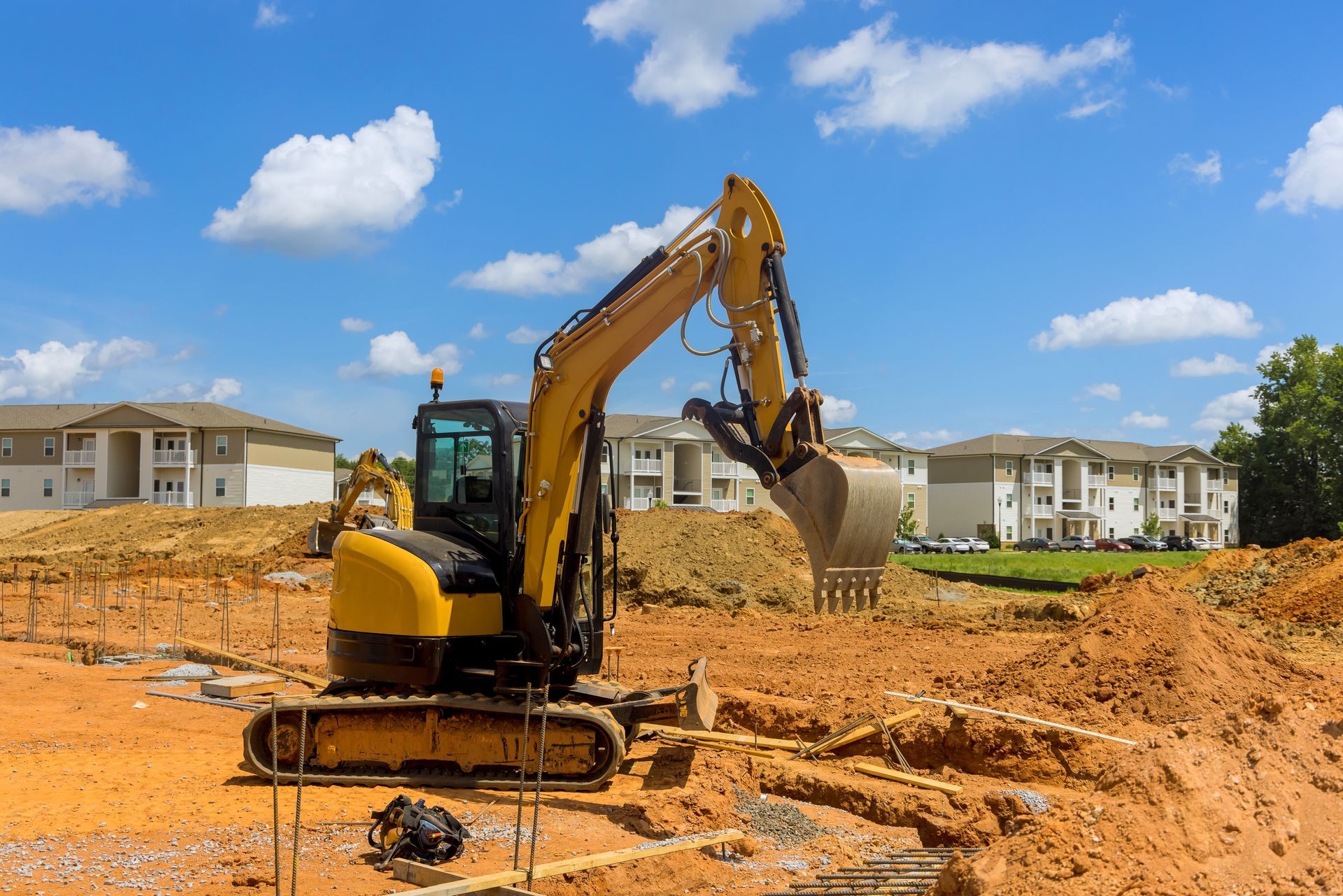 Yellow excavator on a construction site, scooping dirt. Blue sky, white clouds, apartment buildings in background.