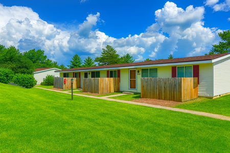 Row of white townhouses with red shutters and brown roofs, set in a grassy yard under a blue sky.