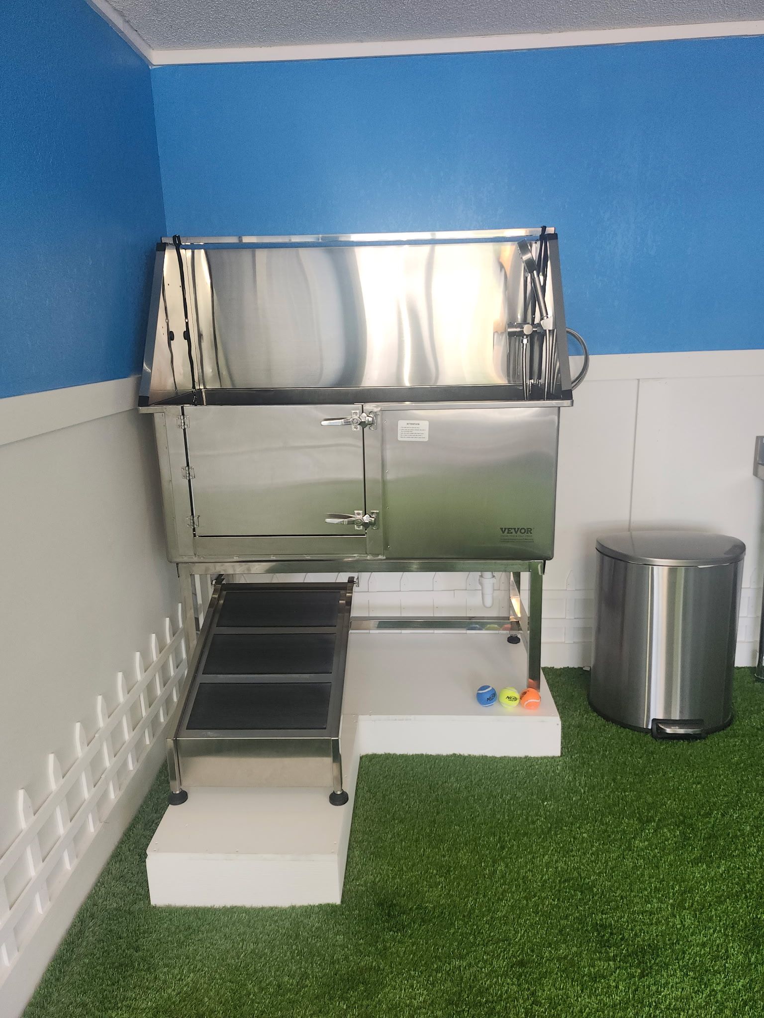 Stainless steel dog grooming tub with stairs, trash can, and accessories, against a blue and white wall.