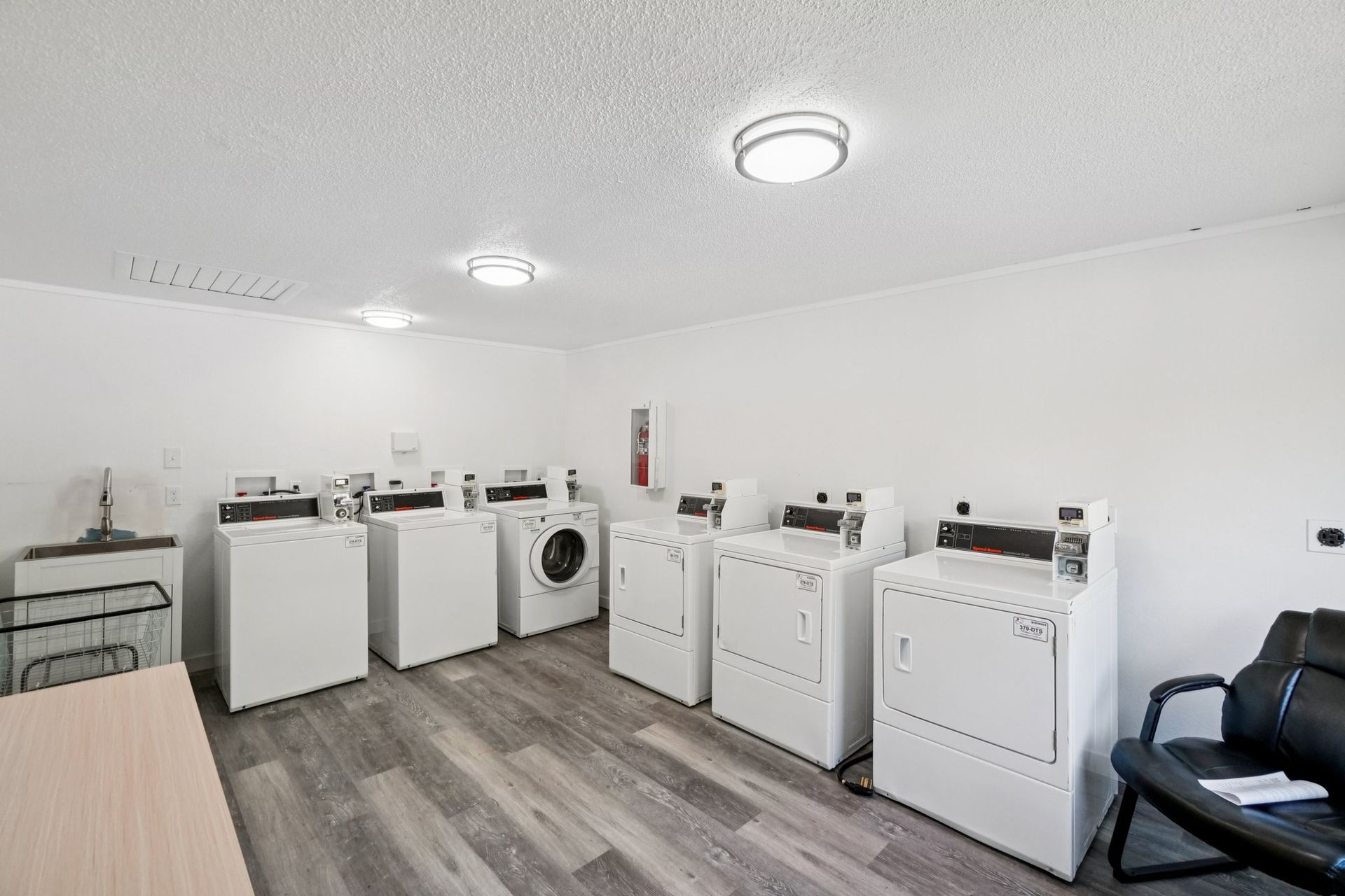 Laundry room with several white washing machines and dryers. Gray flooring, white walls, and a waiting chair.