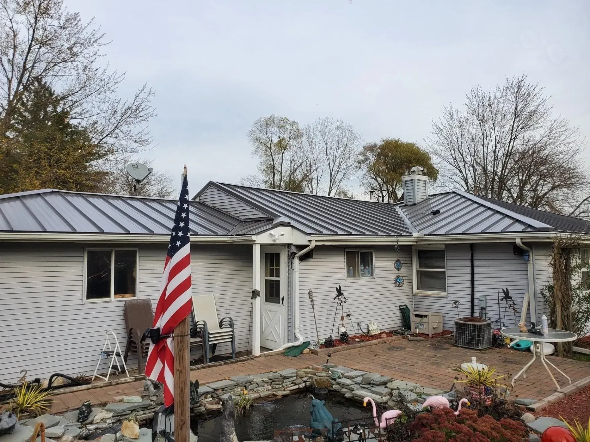 House with gray metal roof, American flag, pond, and cloudy sky.