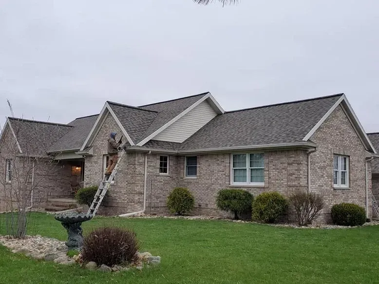 Person on a ladder working on a roof of a brick house on a cloudy day.