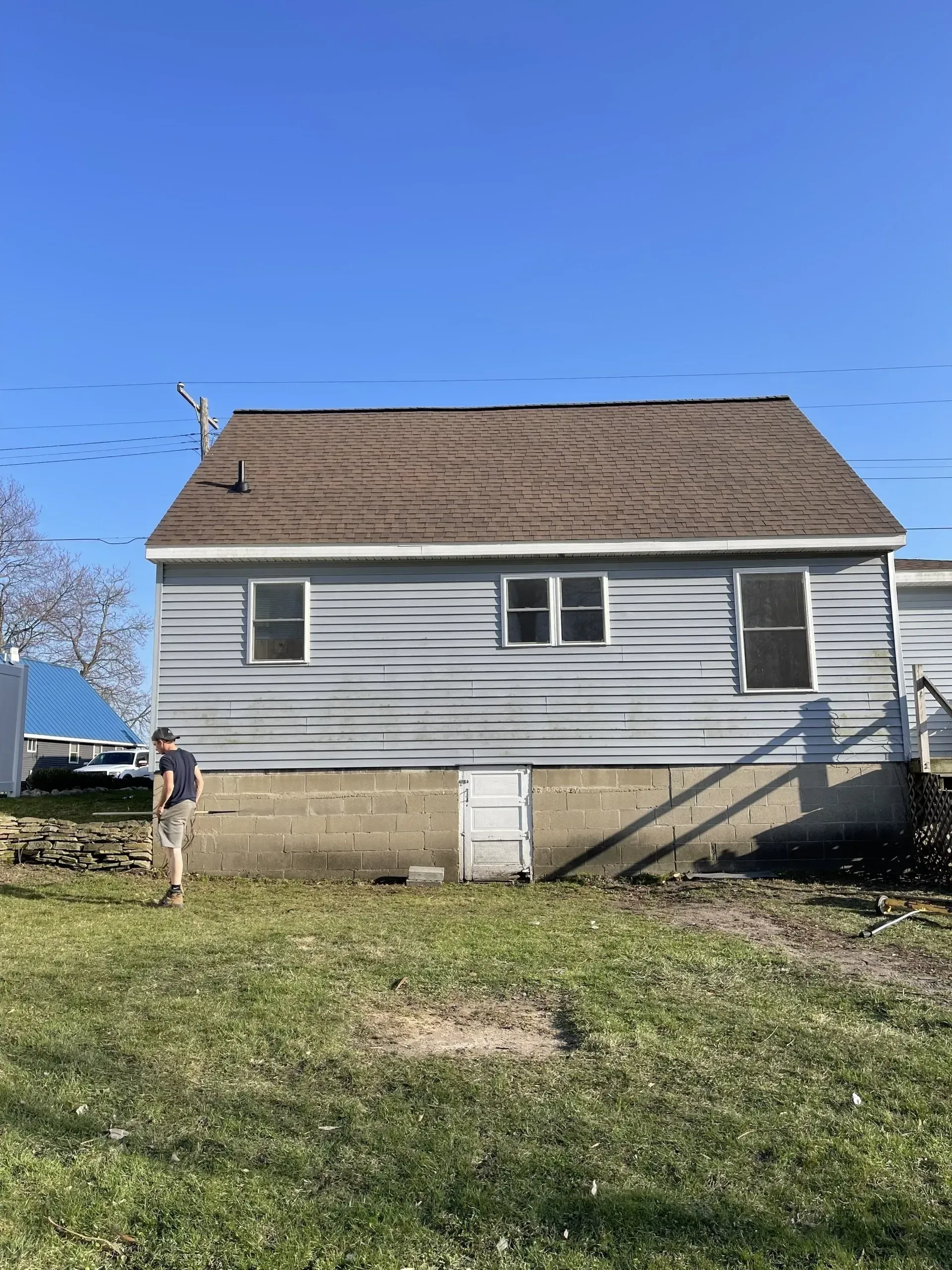 Person digging in front yard of a small house with gray siding and brown roof.