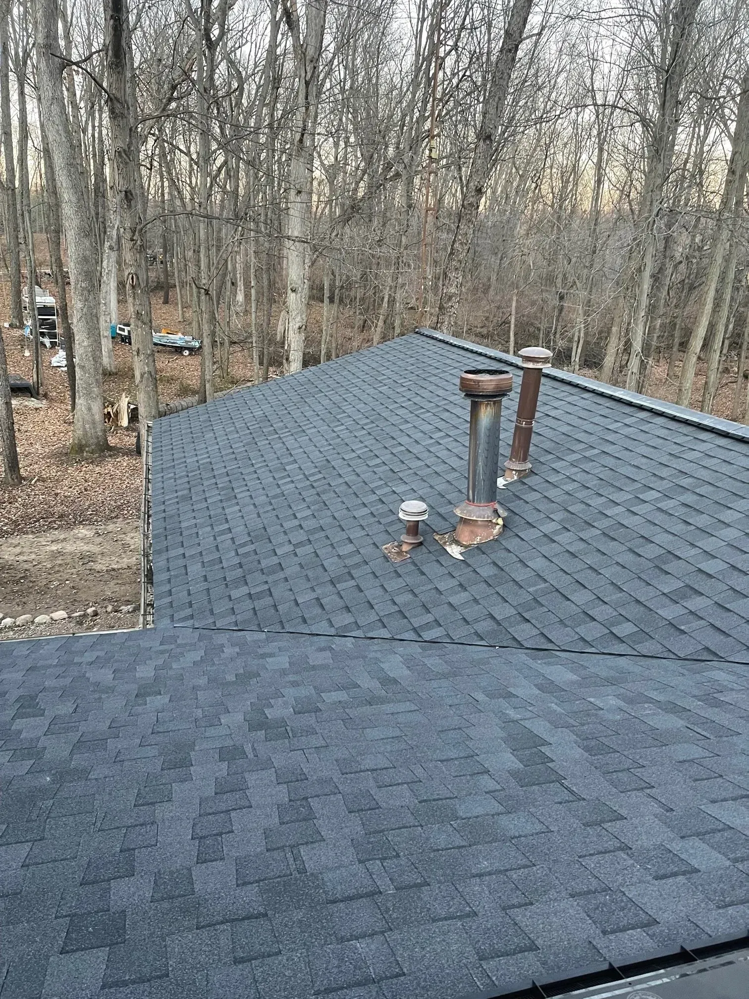 Dark grey shingled roof with two chimneys against a backdrop of bare trees.