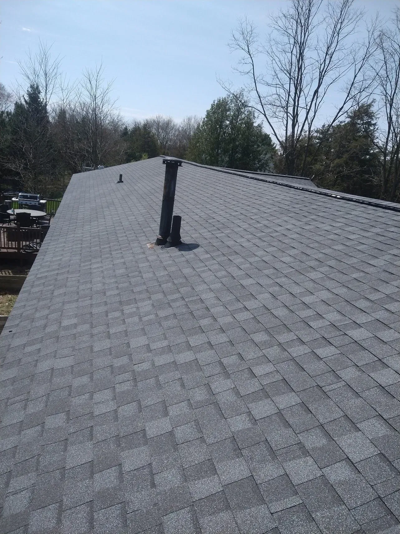 Gray shingle roof with black pipes against a blue sky, surrounded by trees.