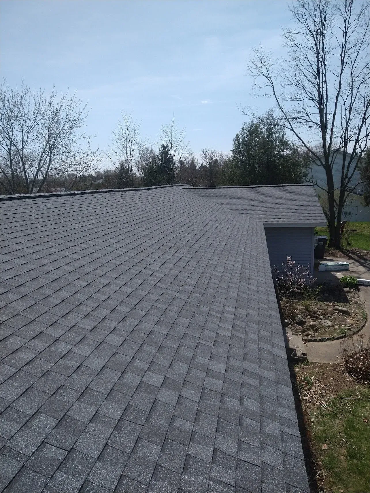 Gray asphalt shingle roof on a house, clear sky, trees in background.
