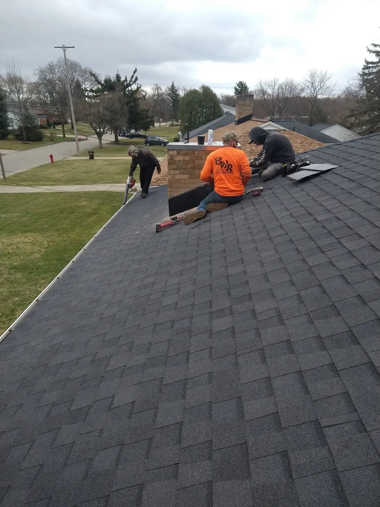 Three workers on a roof, installing shingles near a chimney.