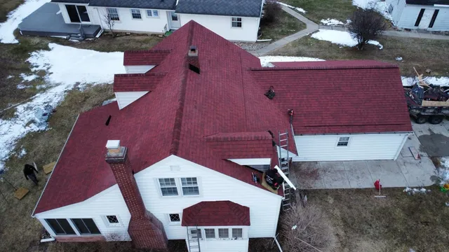 An aerial view of a white house with a new red shingled roof, surrounded by patchily melted snow on the ground.