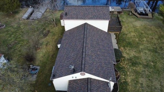 An aerial view of a house with a dark shingled roof, surrounded by a grassy yard near a body of water.