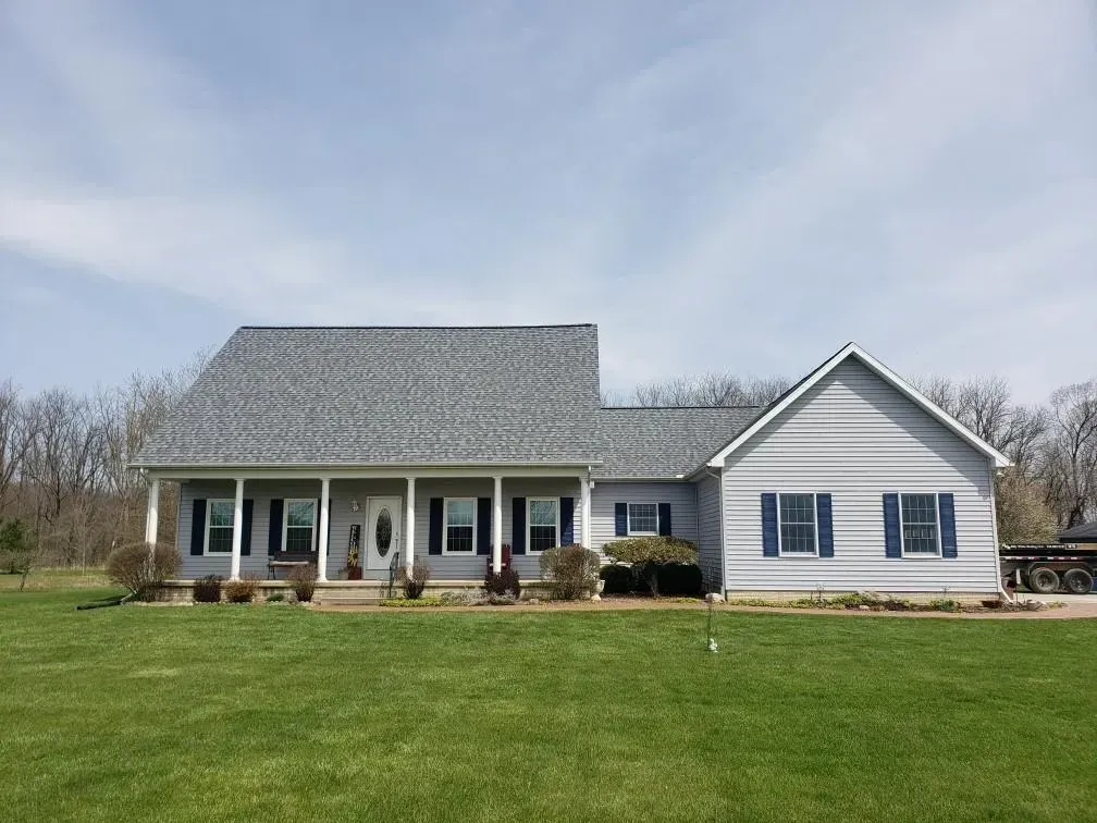 Gray-roofed, light gray house with blue shutters, a porch, and green lawn under a partly cloudy sky.