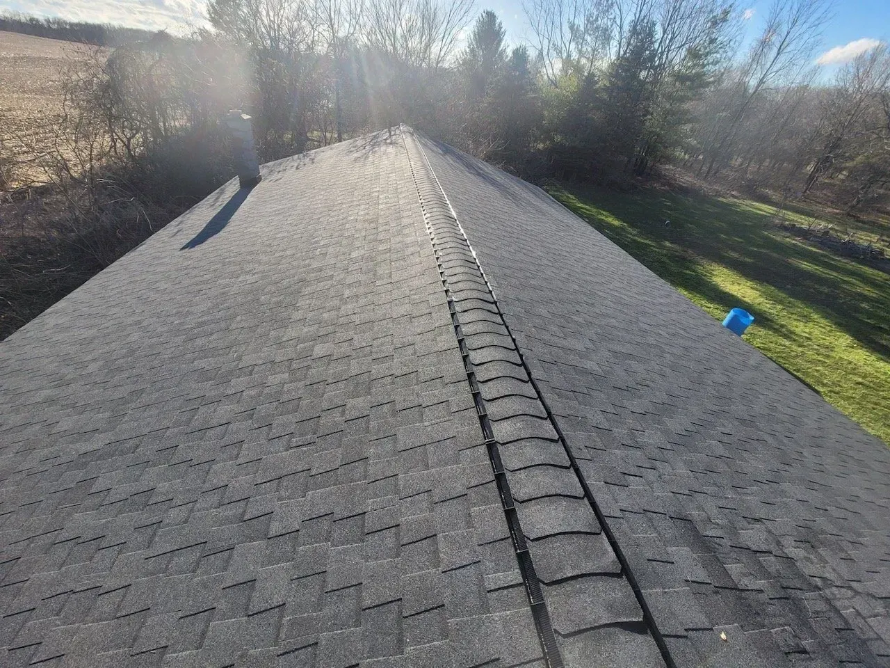 Dark gray shingle roof in sunlight, leading to trees and field. 