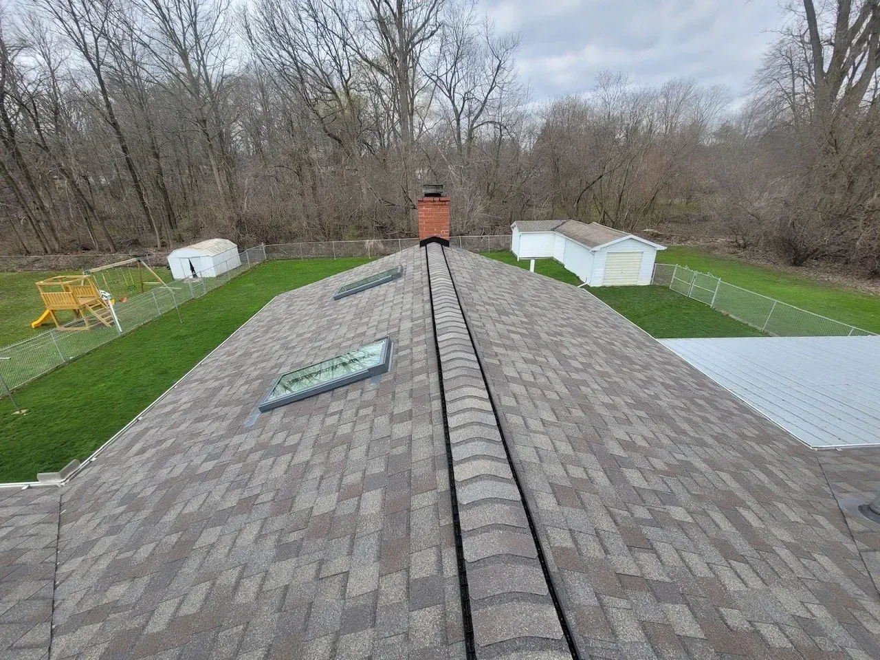 View of a brown shingled roof with a chimney and skylights, green lawn, and trees in background.