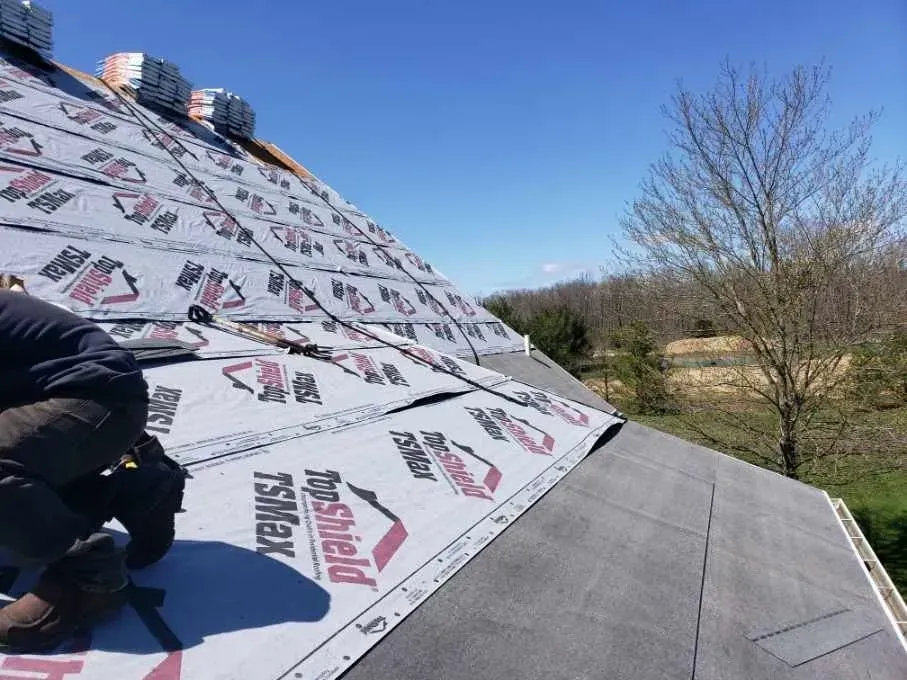 Roofer installing roof underlayment on a house under a blue sky, with shingles stacked nearby.