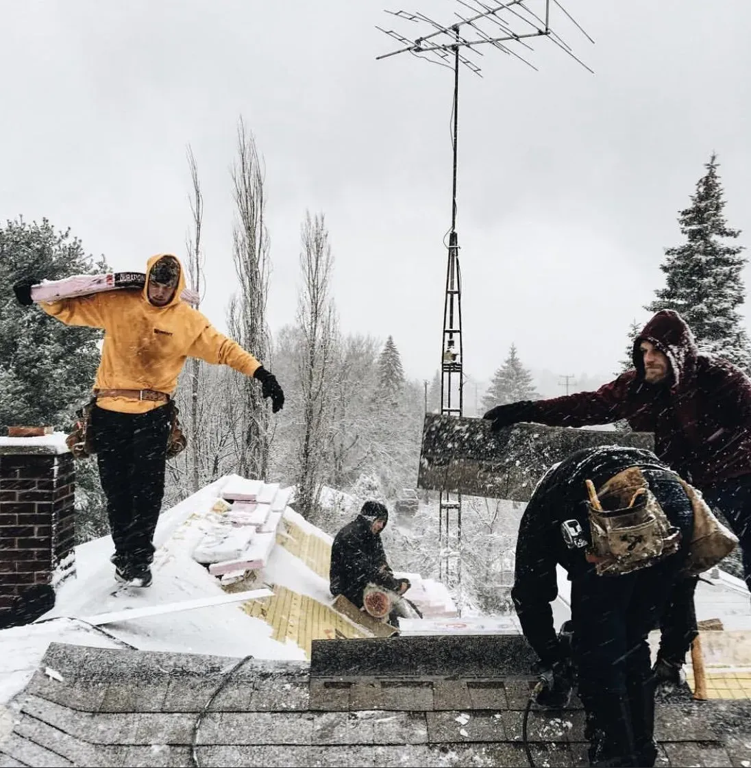 Workers on a snowy rooftop, one carrying insulation, others working on the roof.