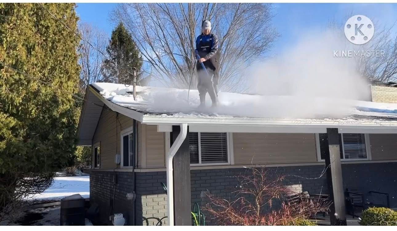 Person on roof using a pressure washer to remove snow from a house on a sunny day.