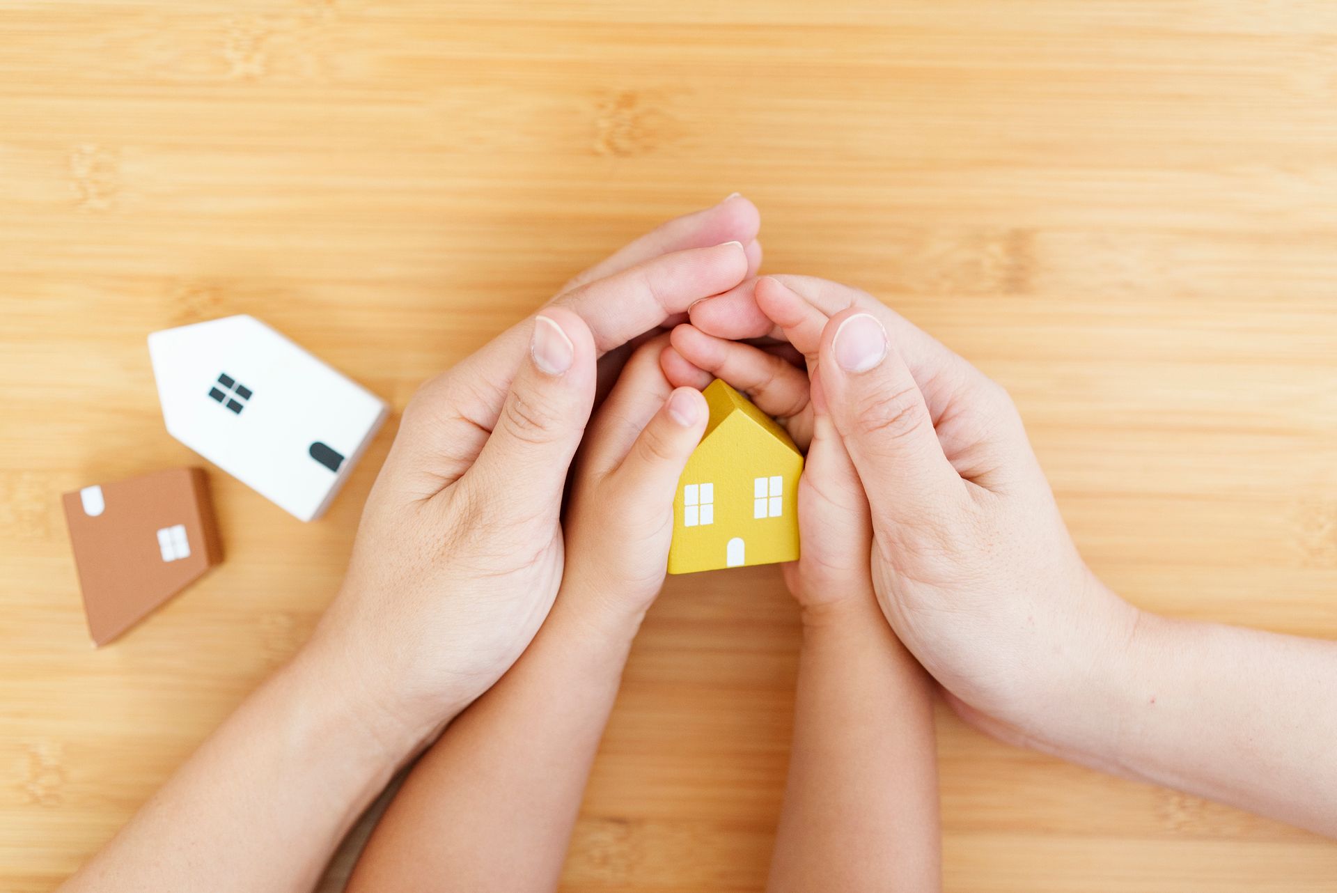 Hands of an adult and child cupping a small yellow house; two other toy houses on a wood surface.