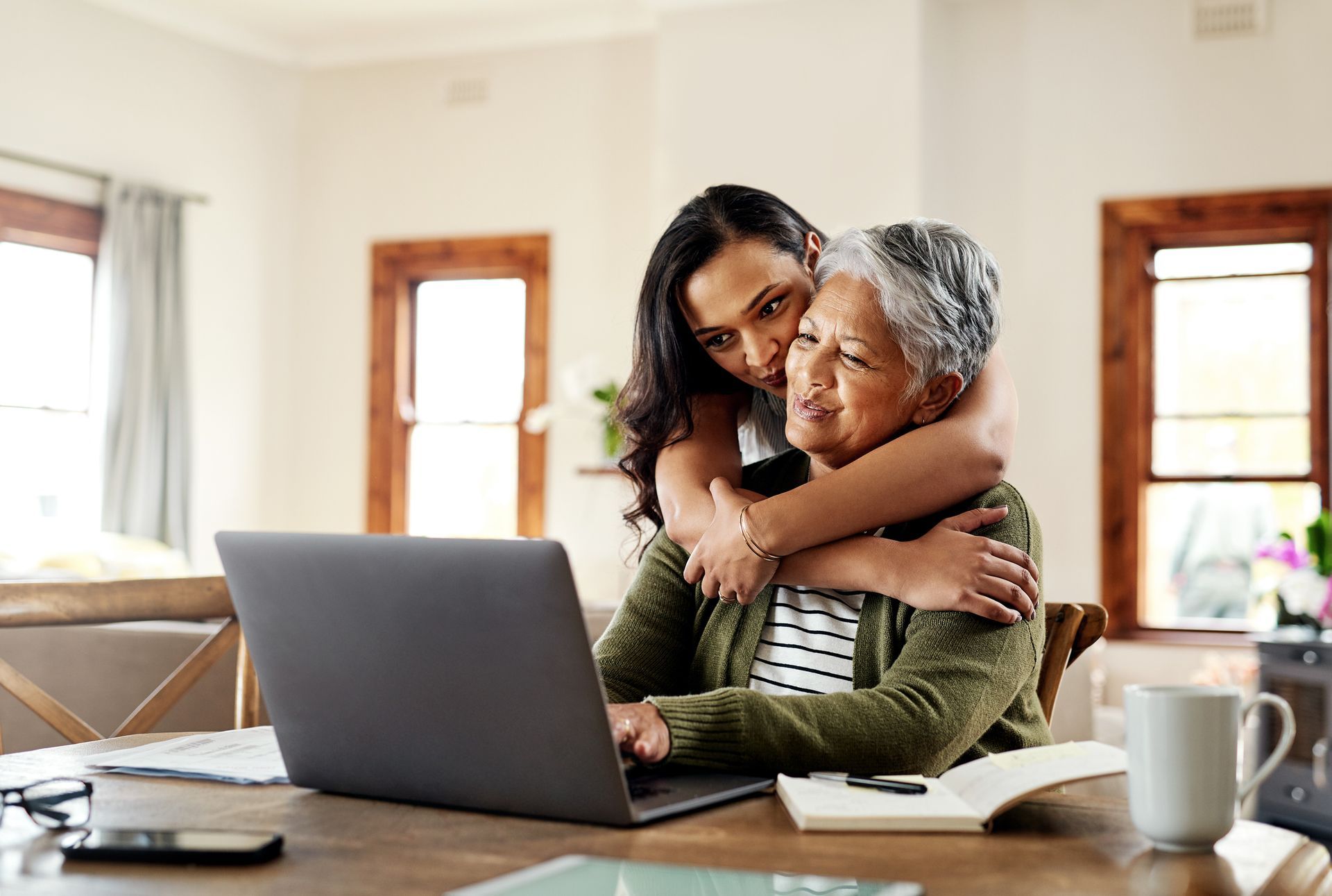 Woman hugs older woman working on laptop at table.