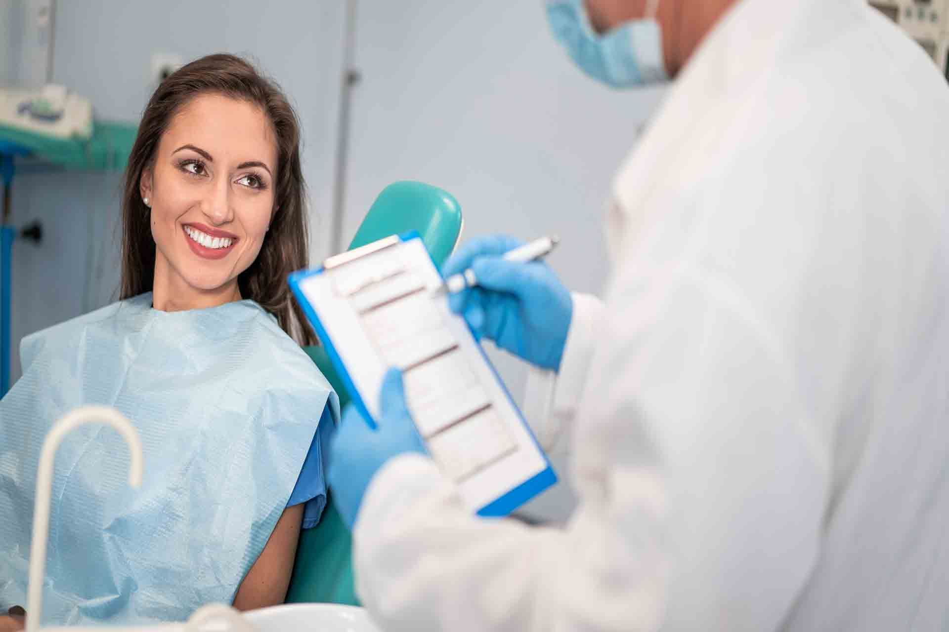 Woman in dental chair smiles at dentist filling out a form. Blue chair and drape.