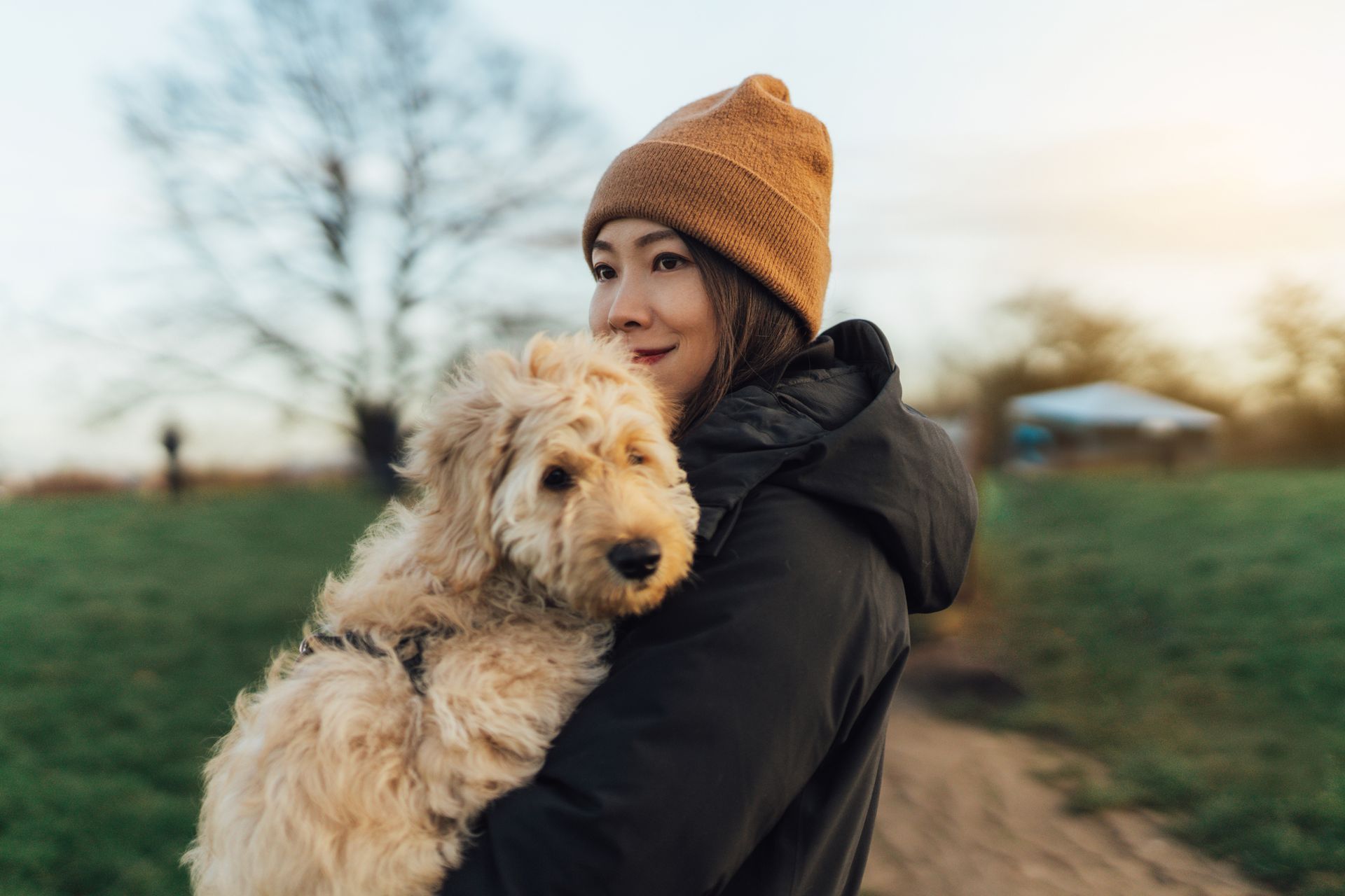 Woman in a brown hat and black coat holding a fluffy, light-colored dog outdoors.