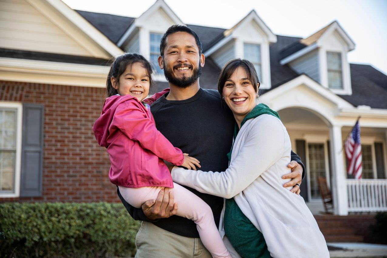 Family of three smiling in front of a house; man holding child, woman with arm around both.