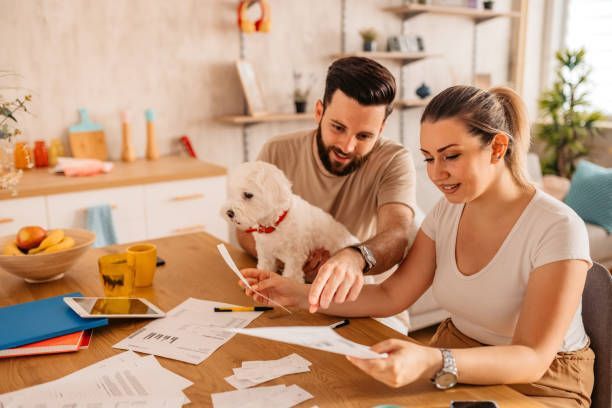 Couple reviews paperwork at a table with a small dog in a light-filled kitchen.