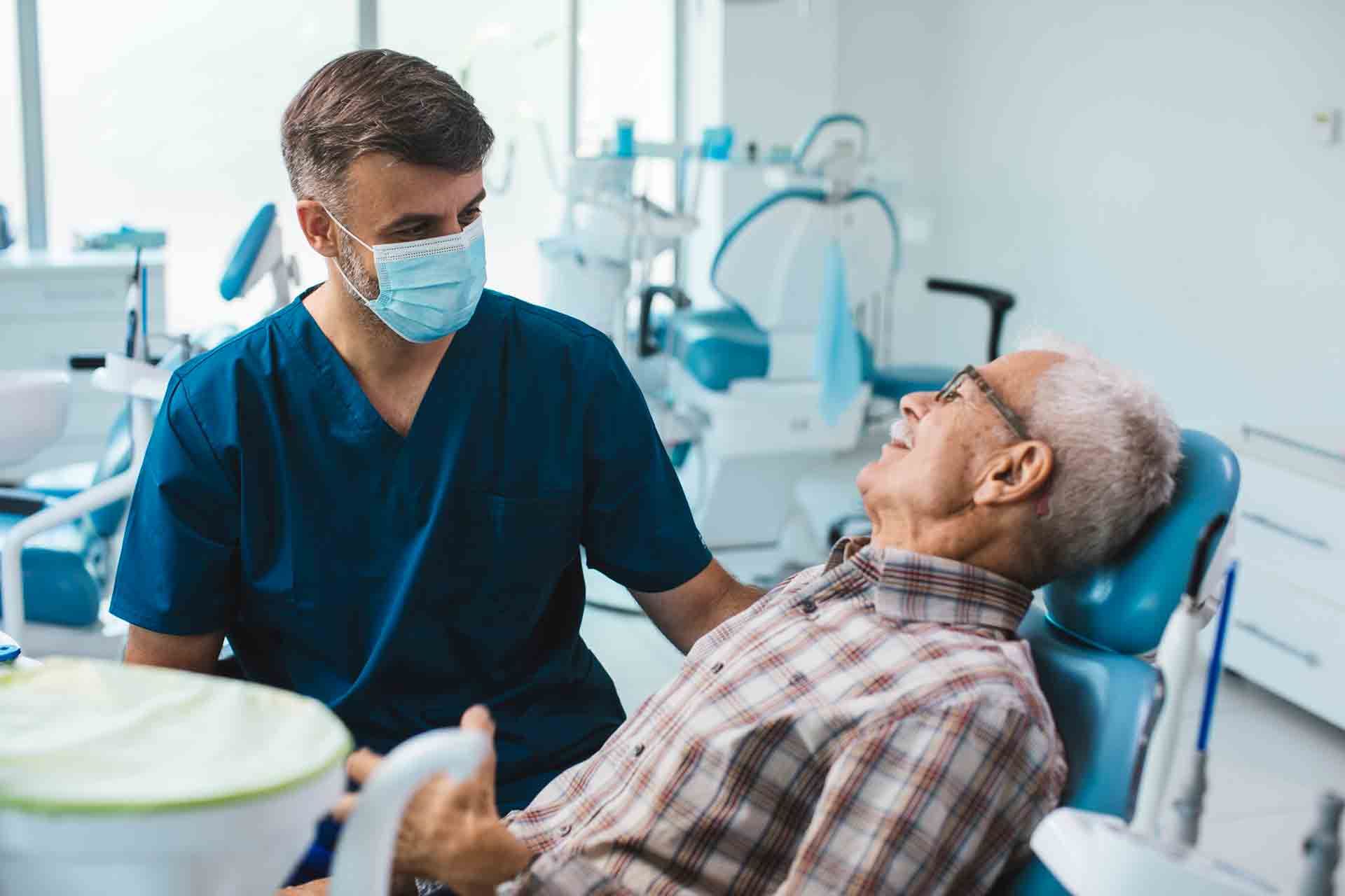 Dentist in blue scrubs and mask with older patient in dental chair, smiling. Clinic setting.