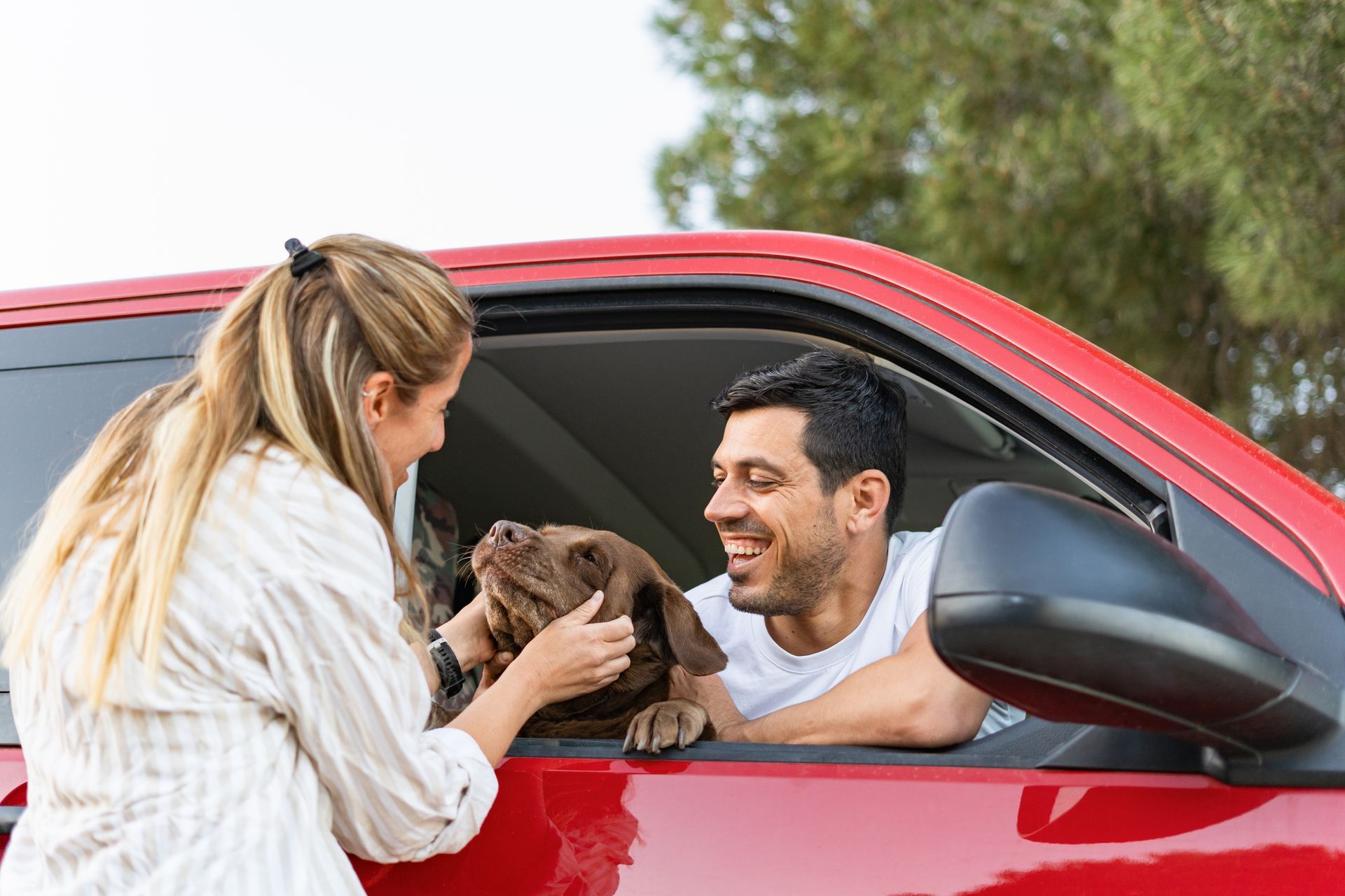 Woman petting a brown dog in a red car window; man smiles, outside.