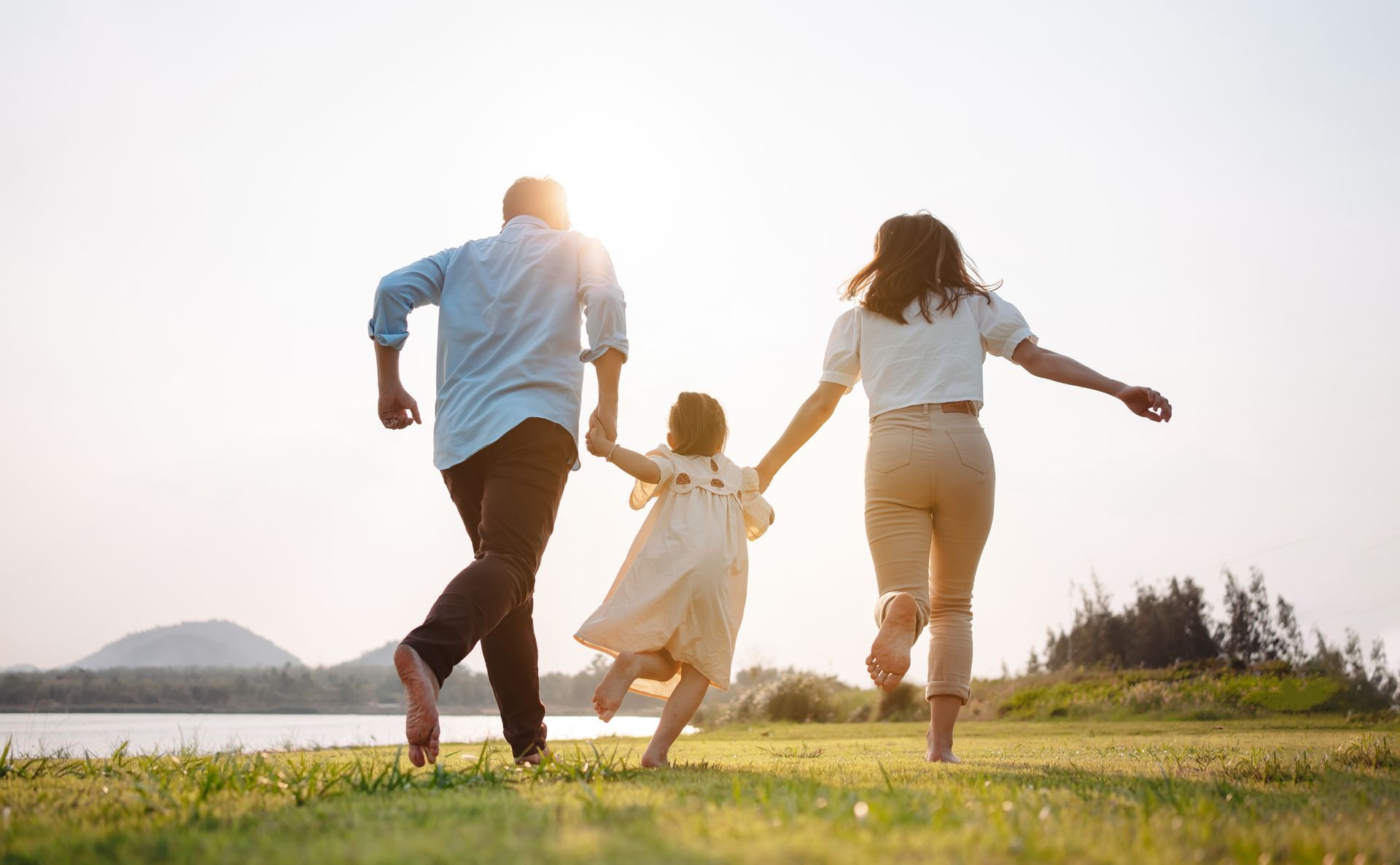 Family runs barefoot on grass towards the water, holding hands, sunny day.