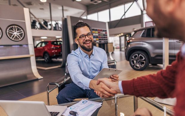 Car salesman shaking hands with a customer in a car dealership; both smiling.