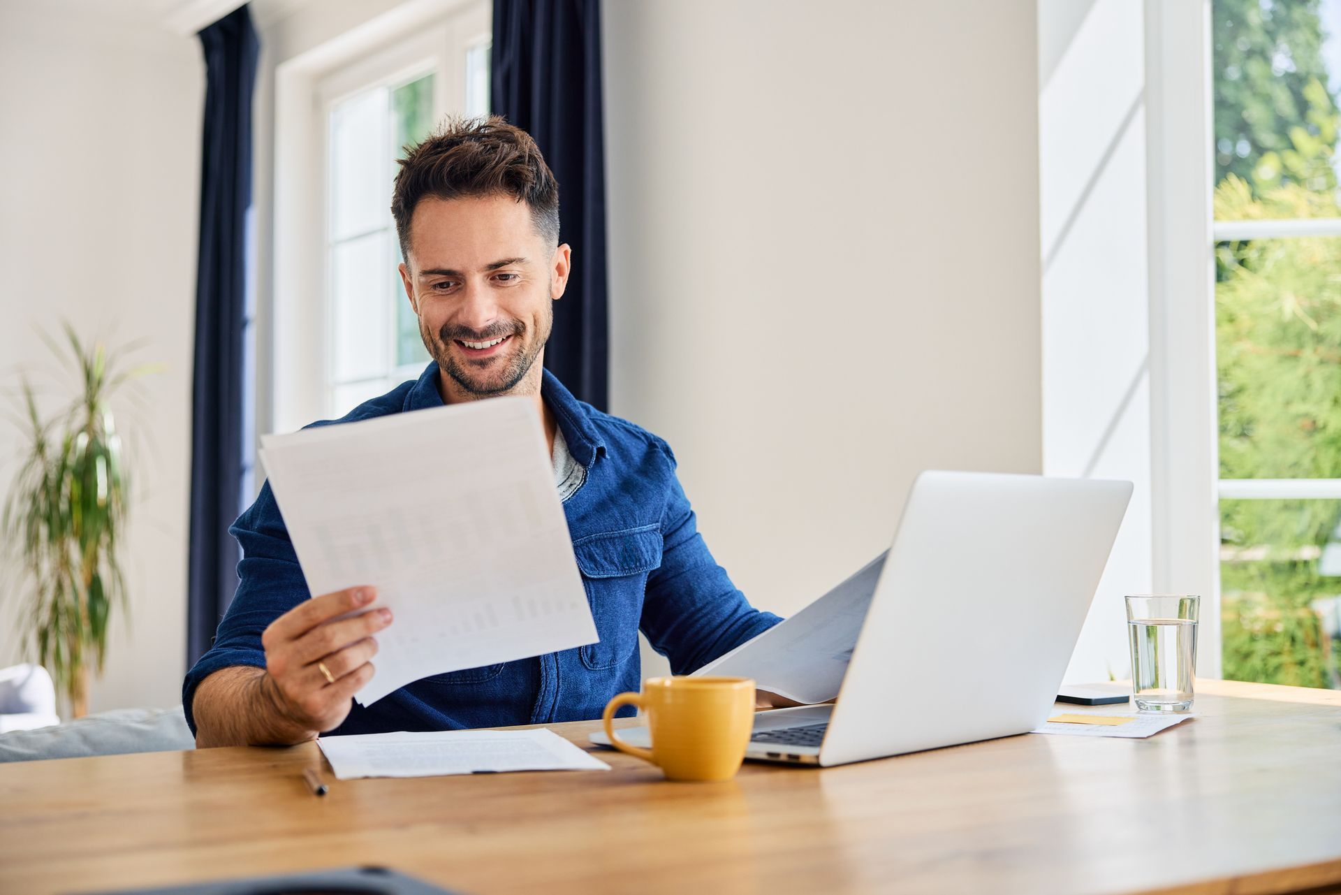 Man smiling while looking at papers and laptop at a desk with a coffee mug and a glass of water, by a window.