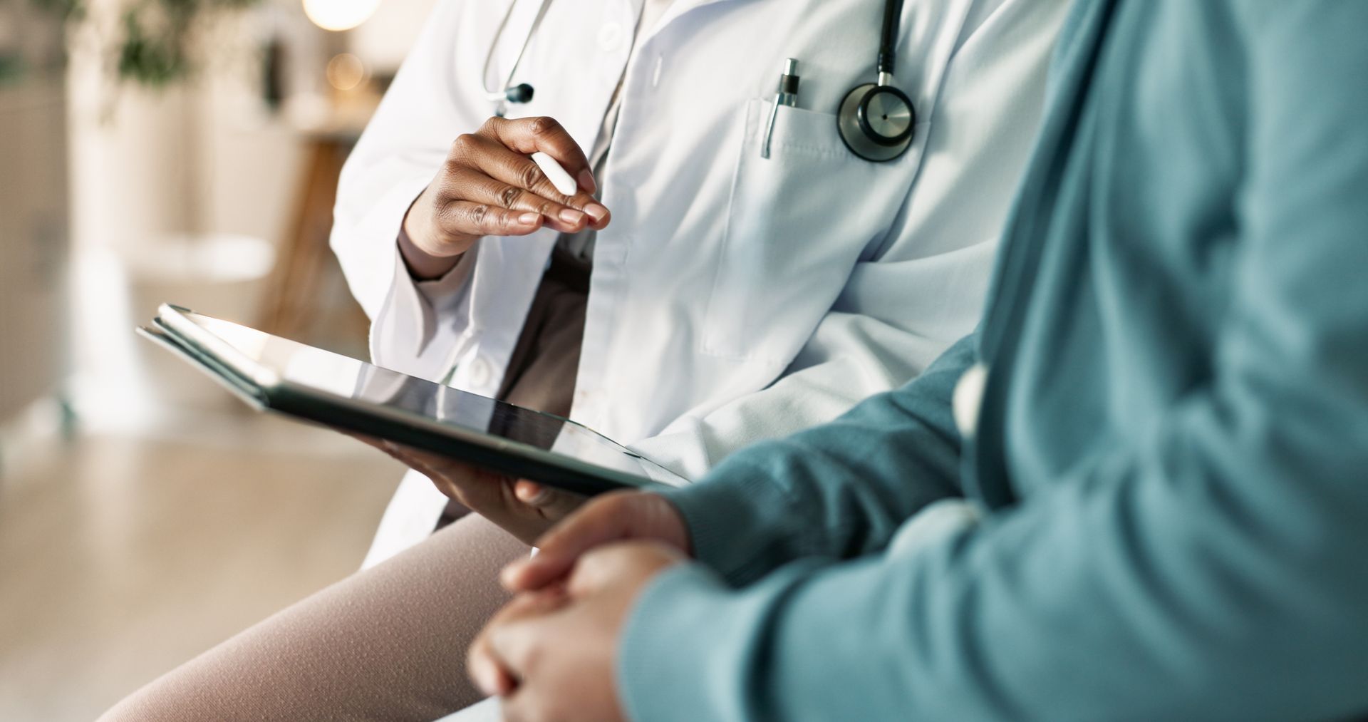 Doctor in a white coat with a stethoscope holding a tablet, consulting with a patient in a blue jacket.