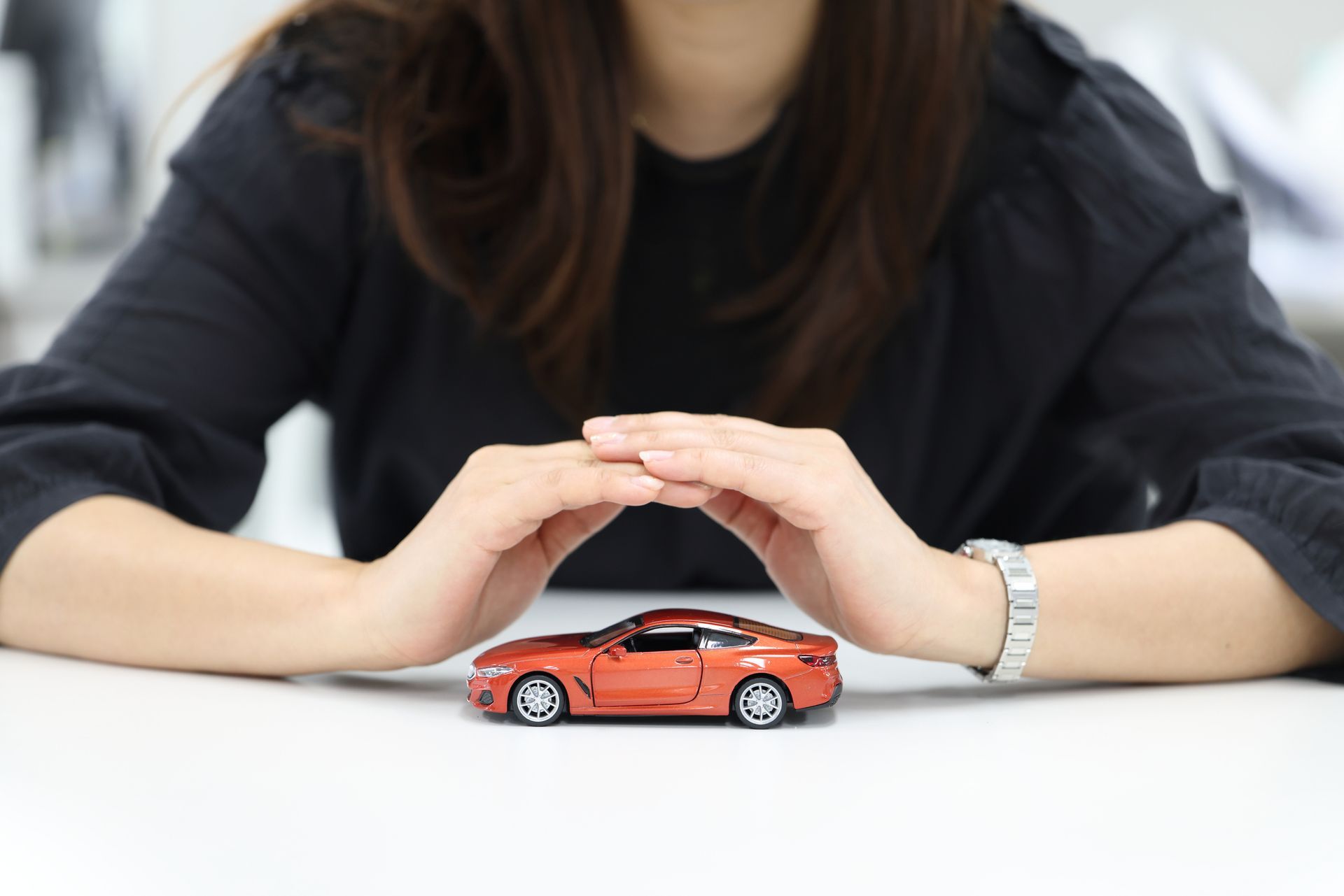 Woman's hands protect a small, orange toy car on a white surface.