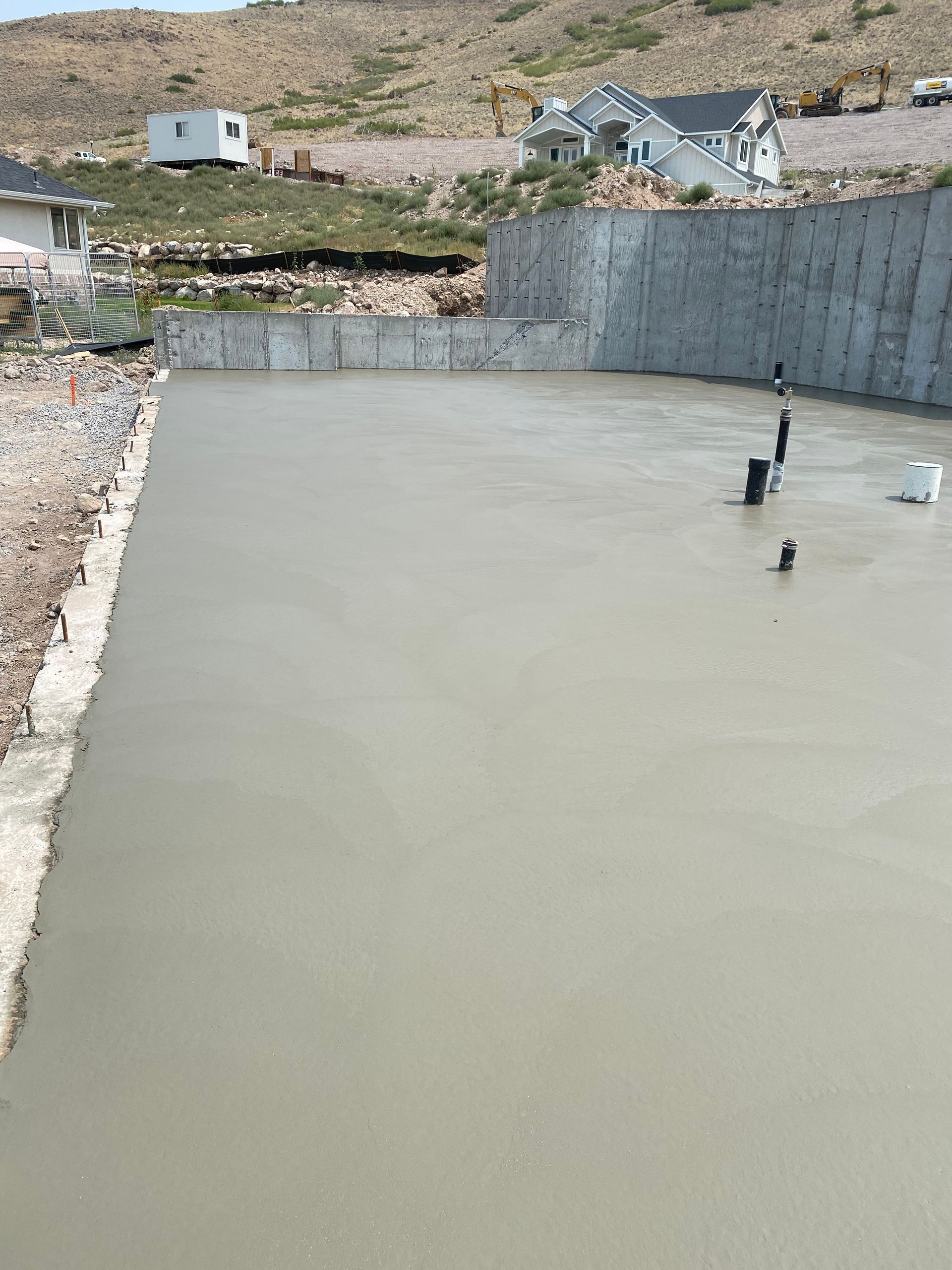 Freshly poured concrete foundation; construction site with retaining wall and hillside backdrop.