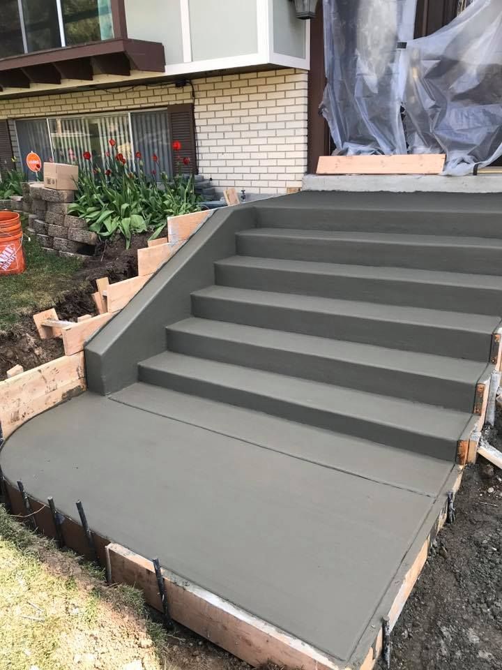 Newly poured gray concrete steps and walkway, bordered by brick and wood forms, leading to a house entrance.