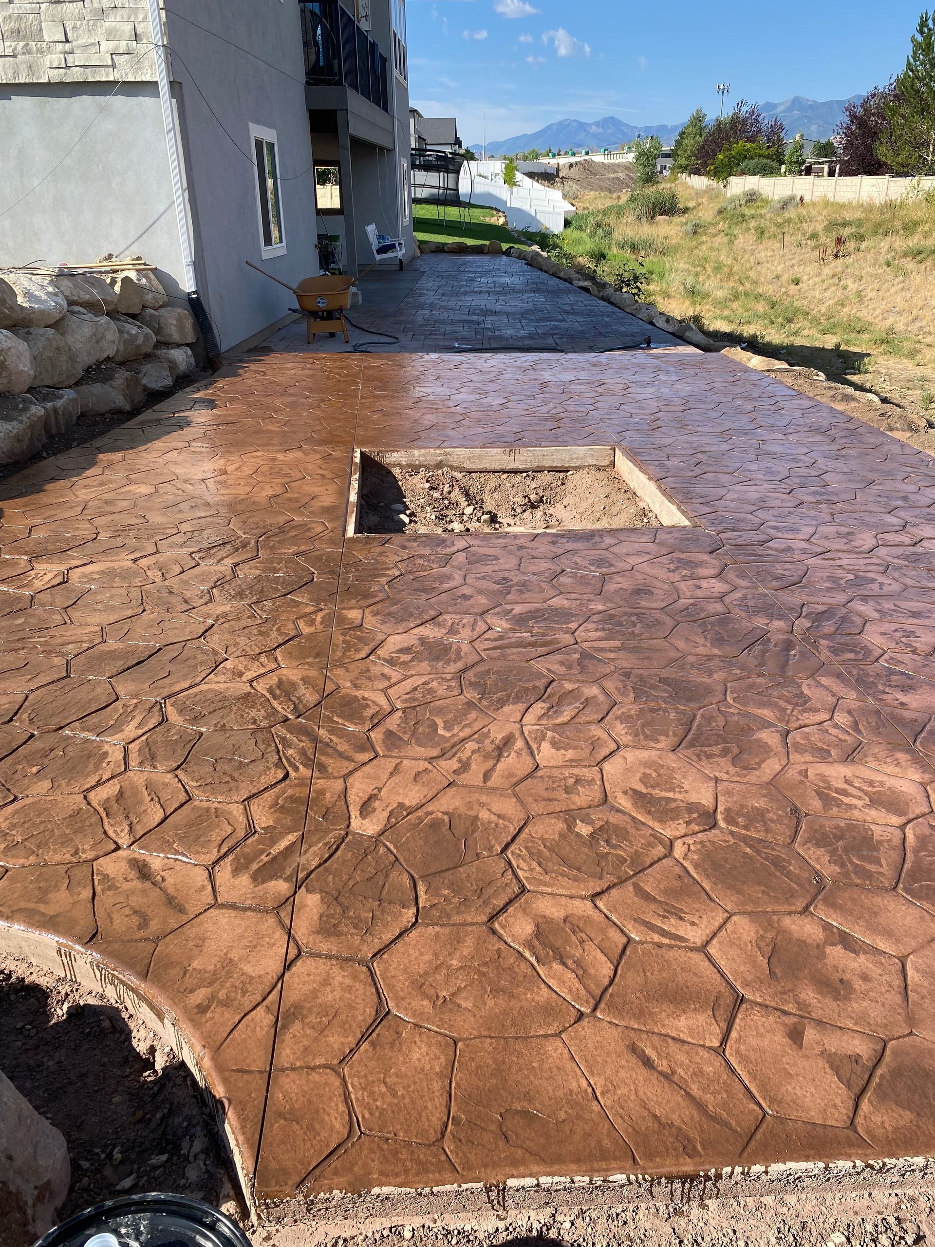 Brown stamped concrete patio with fire pit opening, next to a gray house, with a grass area.