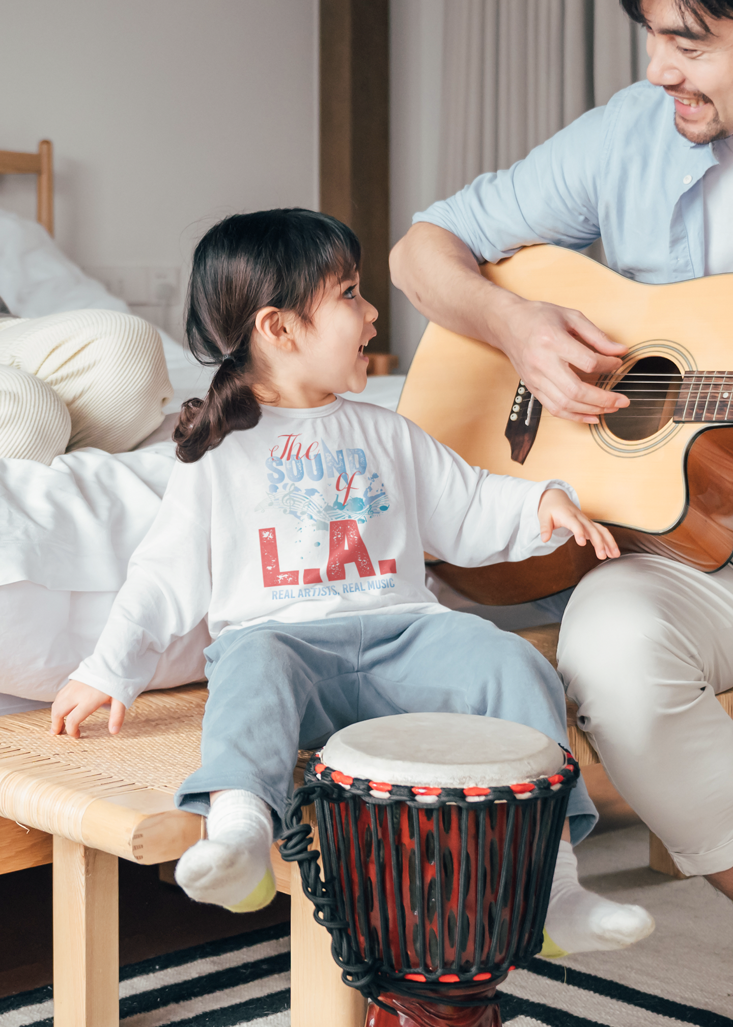 A man is playing a guitar and a little girl is sitting on a drum.