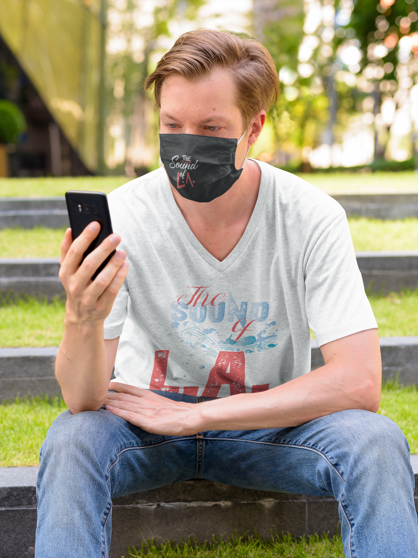 A man wearing a face mask is sitting on steps looking at his cell phone.