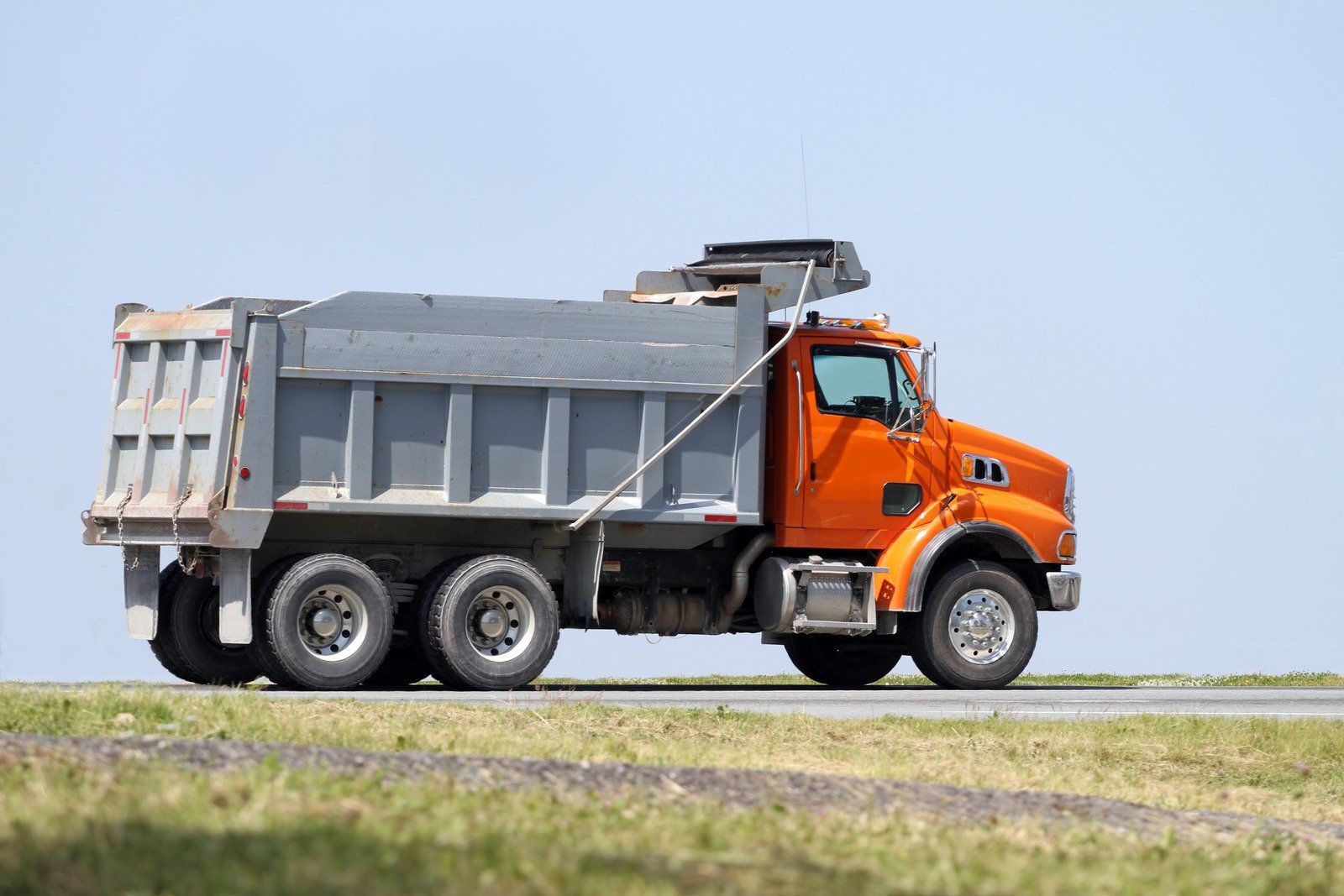 Orange dump truck on a road, gray bed raised, clear blue sky in the background.